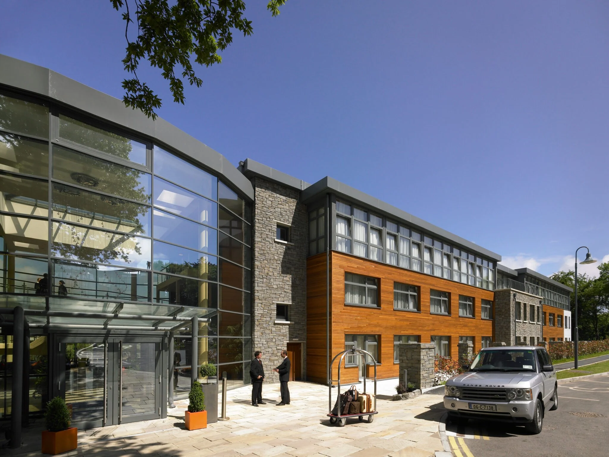 Modern building with large glass windows and wooden paneling, two people talking outside, parked Range Rover, luggage cart, sunny day with blue sky.