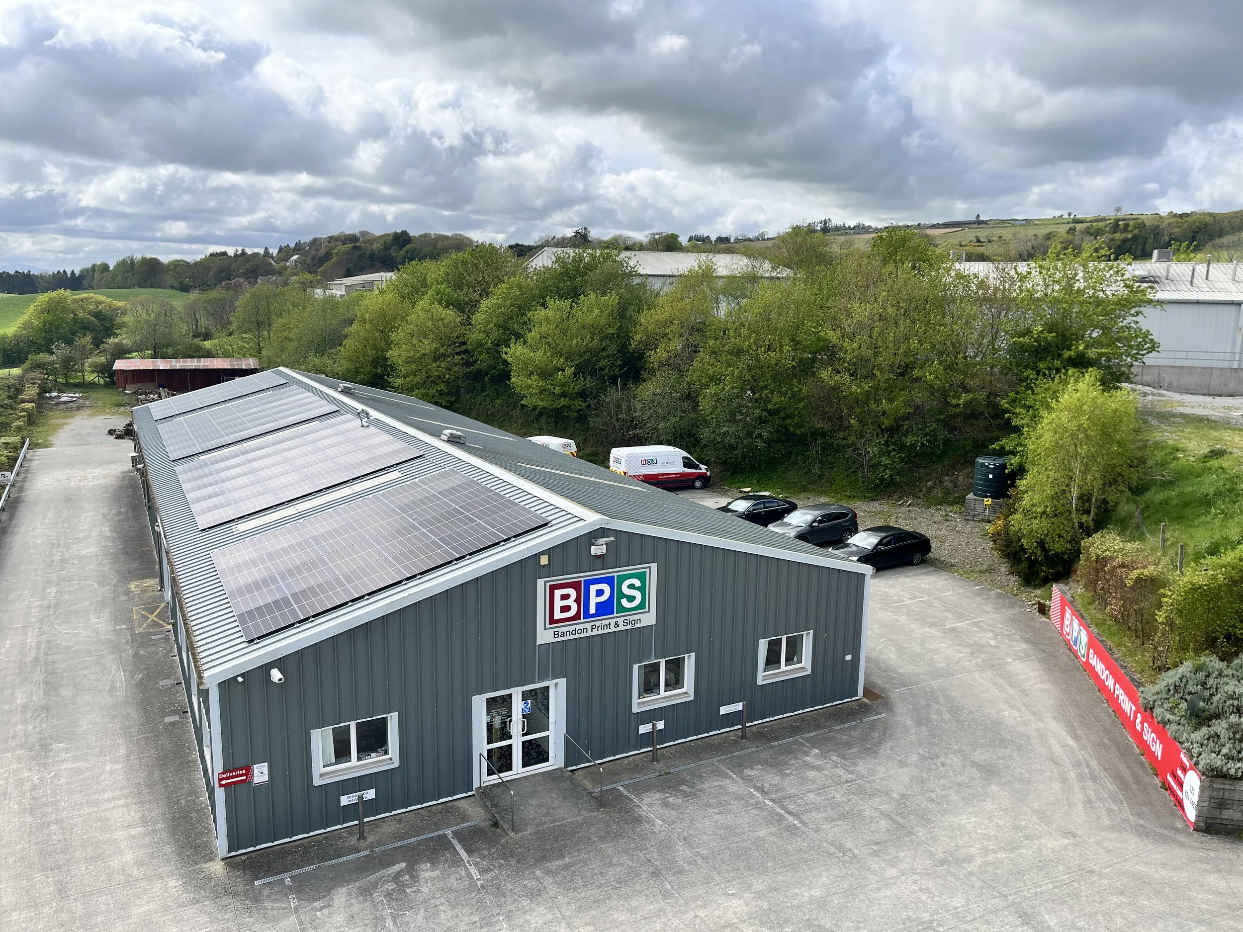 A commercial building with large solar panels on the roof, labeled Bandon Print & Sign, with parking lot and greenery in the background.