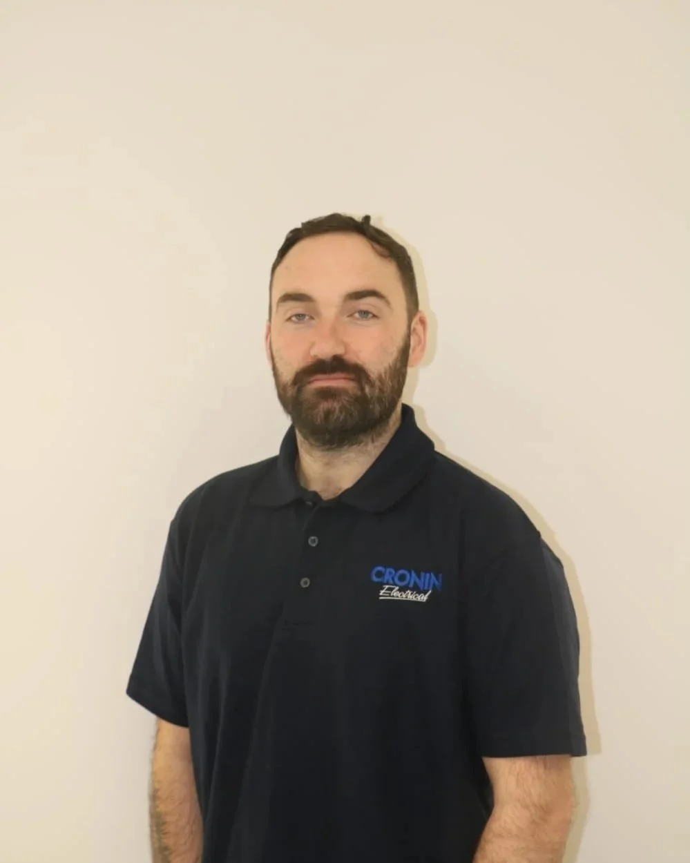 Man with dark hair and beard wearing a black polo shirt with 'CRONIN Electric' logo, standing against a plain white wall.