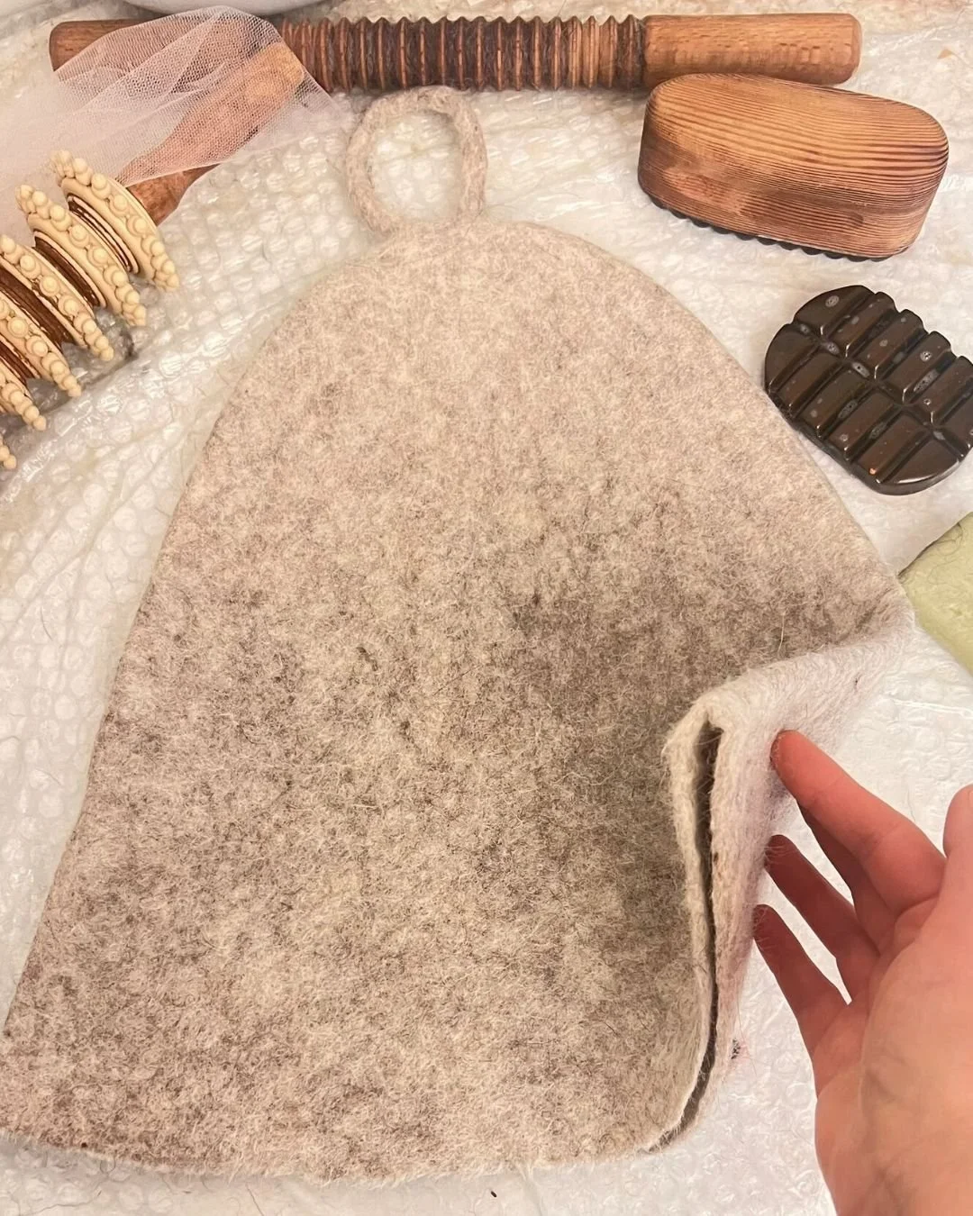 Dovile's hand showing the dense undyed Tyrolean wool of a Feltforma sauna hat in her Vilnius studio — surrounded by hand-felting tools, water-washed only, no dyes, no chemical treatments, made for sauna and cold plunge rituals