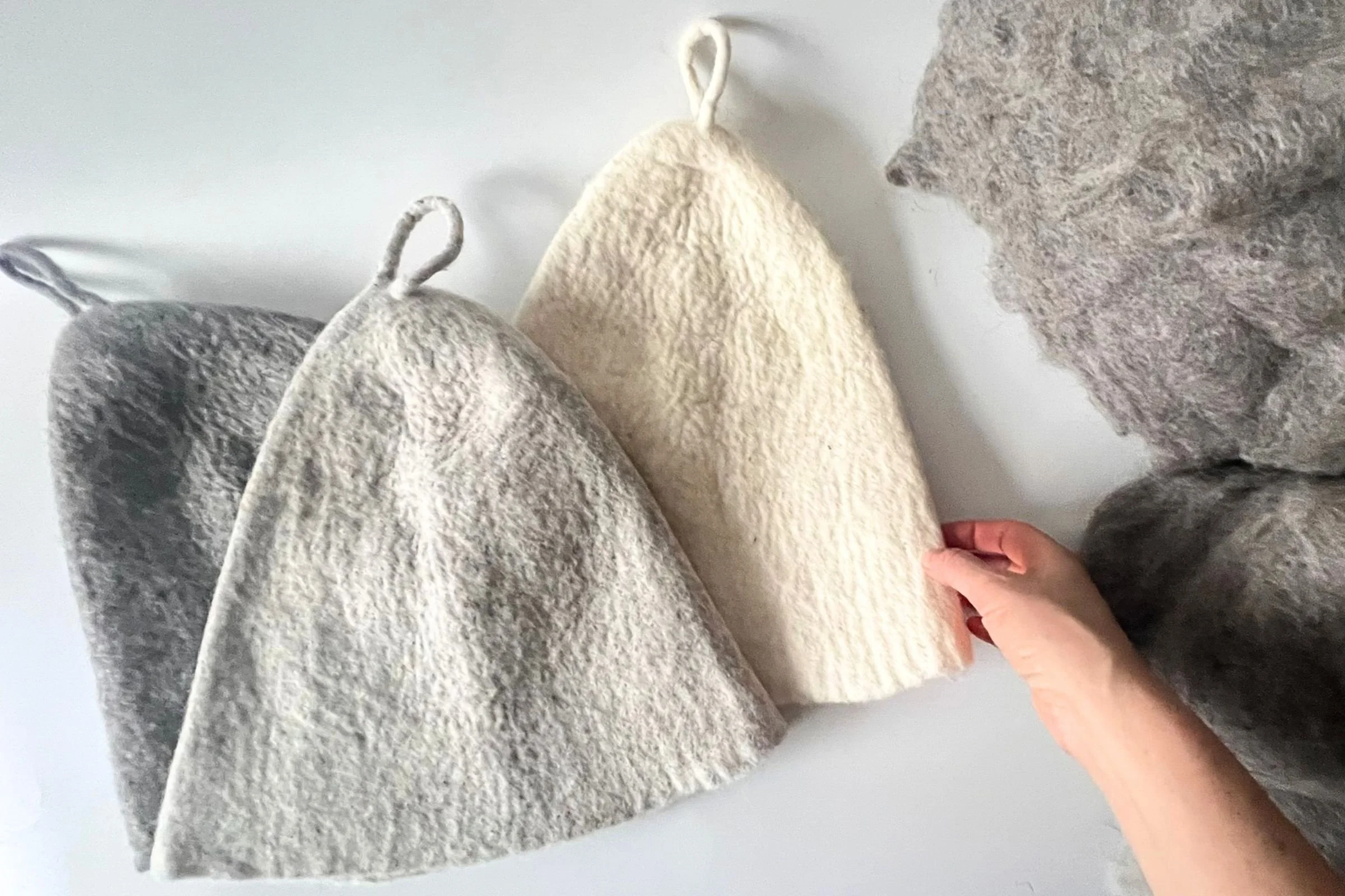 Undyed wool sauna hats held by hand, showing flexible felt structure used during transitions between heat and cooling in sauna rituals