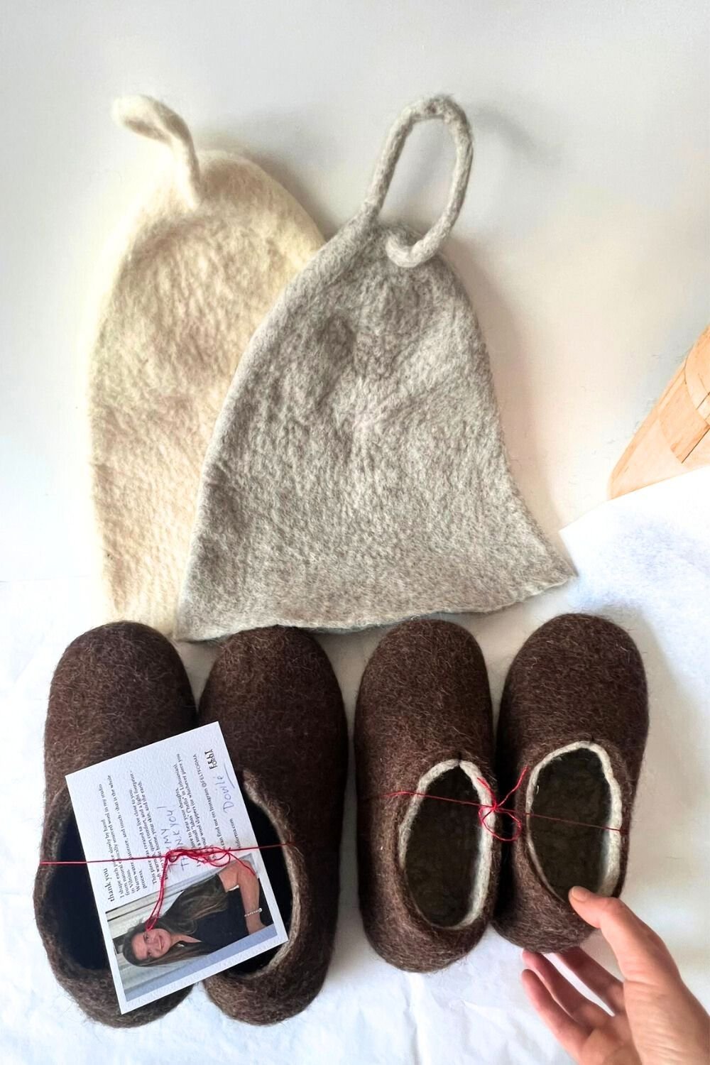 Two undyed Feltforma wool sauna hats in natural white and grey tones alongside a pair of dark brown hand-felted wool slippers — complete sauna and cold plunge ritual set, handmade in Vilnius, Lithuania