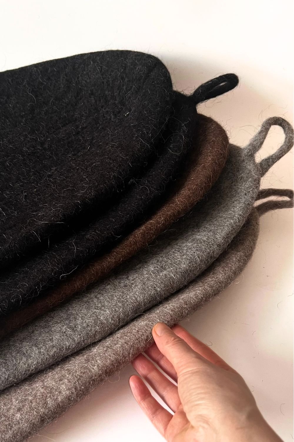 Five handmade undyed wool sauna hats stacked — natural charcoal, brown, grey and light grey Tyrolean mountain wool. Dense, seamless construction for infrared sauna, contrast therapy and cold plunge. Made in Vilnius, Lithuania.