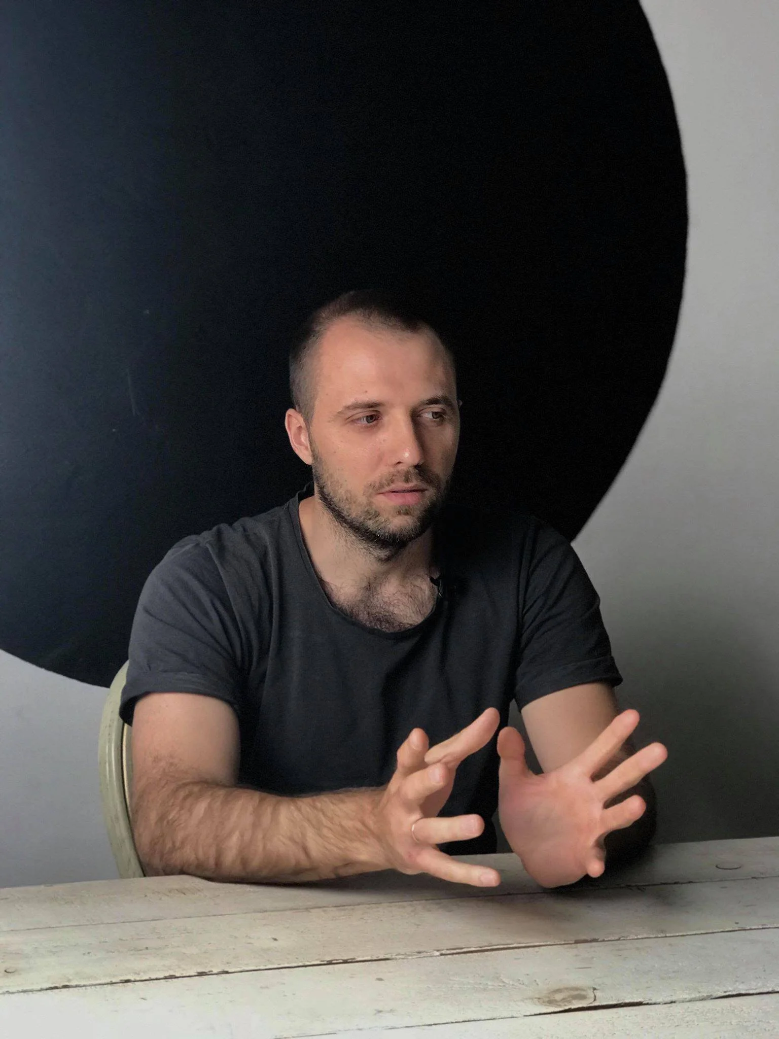 A man with short dark hair and a beard, wearing a black t-shirt, is sitting at a wooden table with a serious expression, gesturing with his hands, in front of a background featuring large black and white shapes.