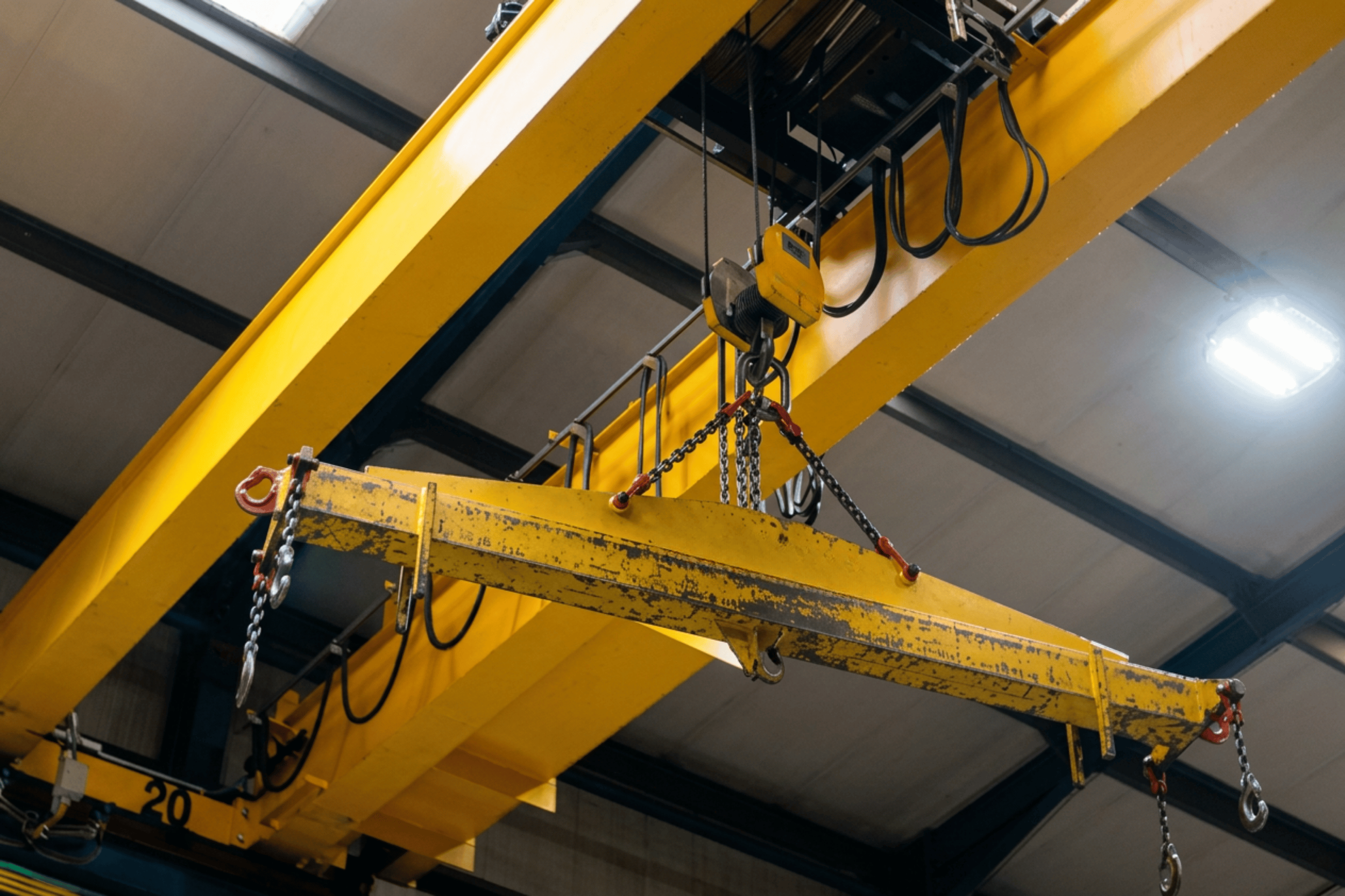 Close-up of a yellow overhead crane with hooks and chains, inside an industrial warehouse.