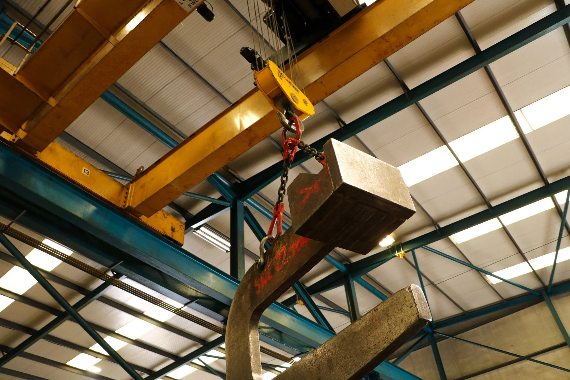 A large industrial hook lifting a concrete block in a warehouse with metal beams and skylights.