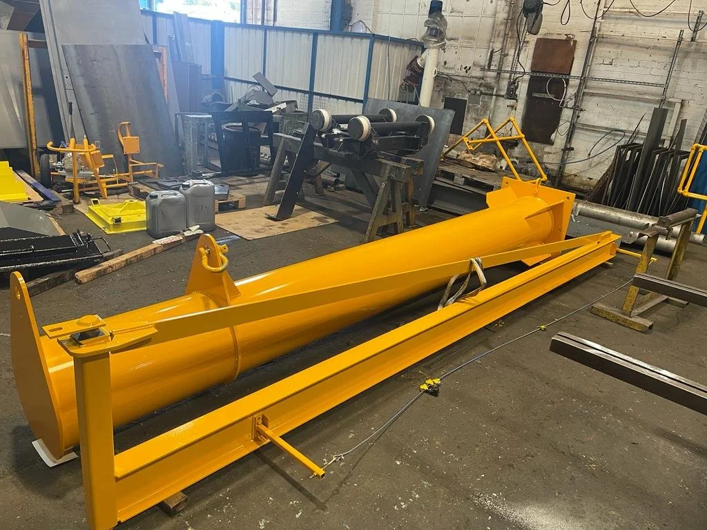 A large yellow industrial snow plow blade in a workshop or warehouse with metal tools and equipment around it.