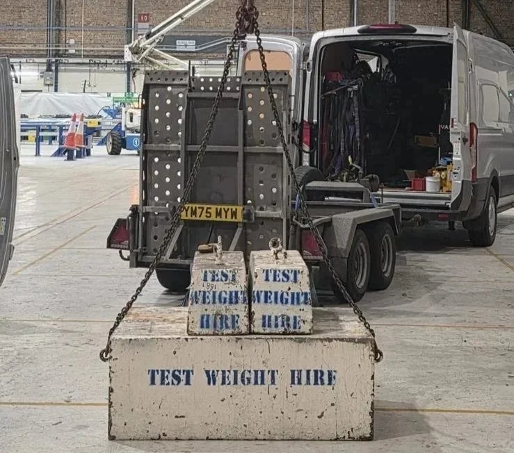 A vehicle with a testing weight lift attached to a crane, featuring concrete blocks marked 'TEST WEIGHT HIRE' stacked on a pallet, in an indoor industrial setting.