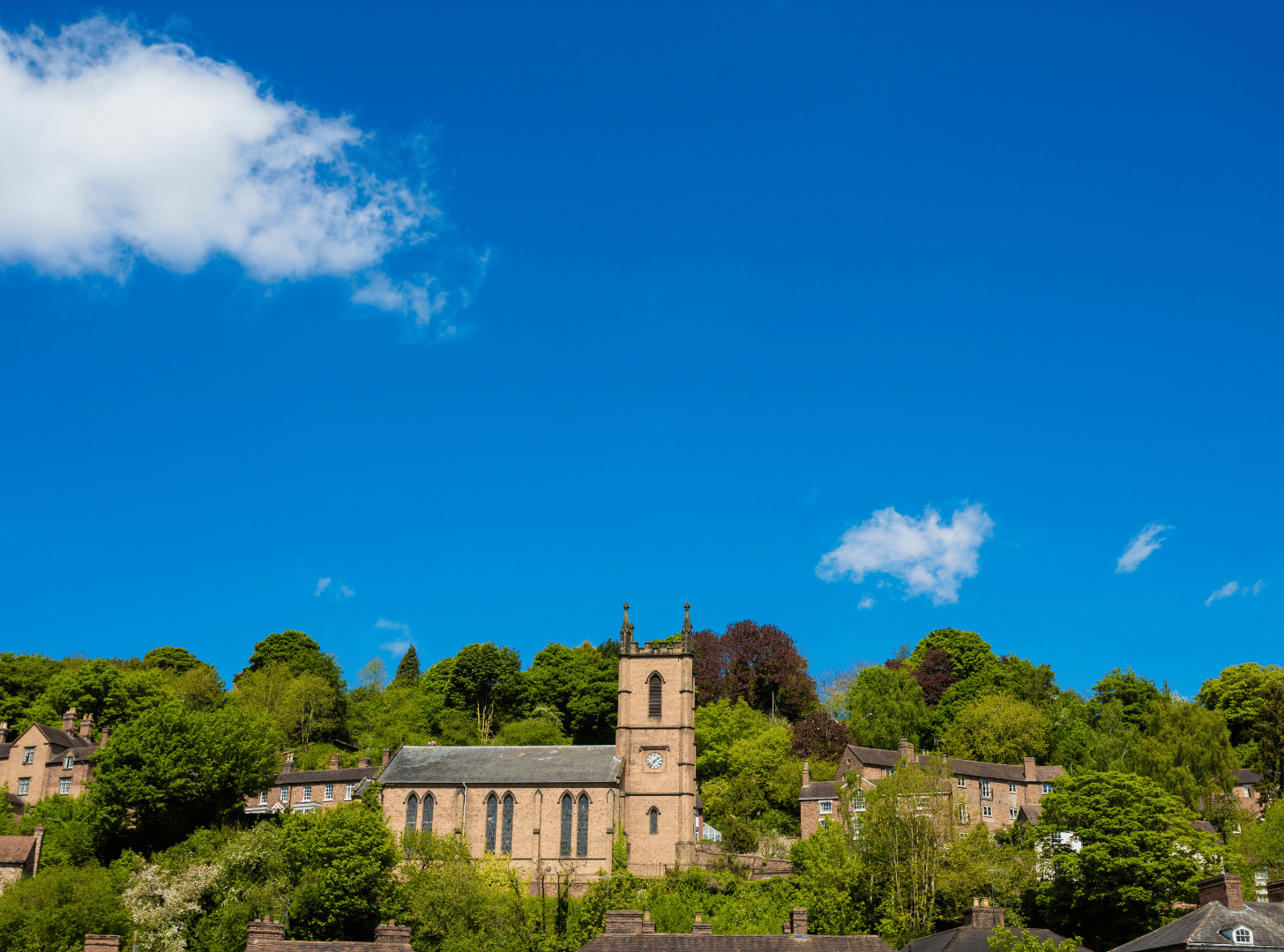 A church with a clock tower situated on a hill, surrounded by trees and residential houses, under a blue sky with a few clouds.