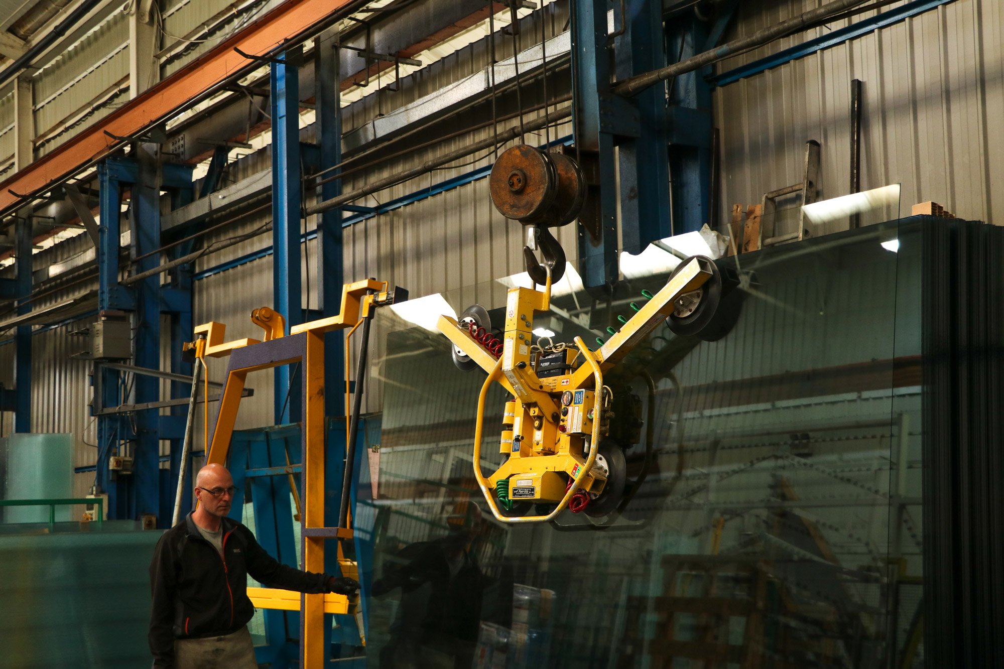 A worker in safety glasses and a jacket operates a yellow robotic machine that handles large glass panels inside a factory.