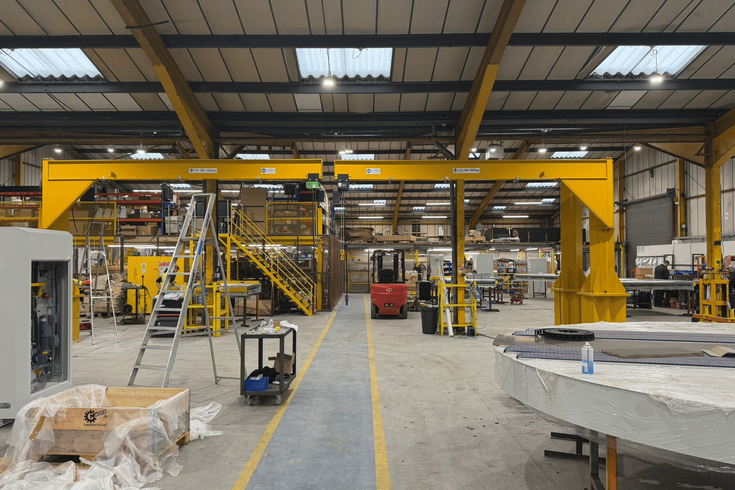 Interior of a large industrial warehouse with yellow overhead cranes, workstations, ladders, tools, and equipment in progress of assembly or manufacturing.