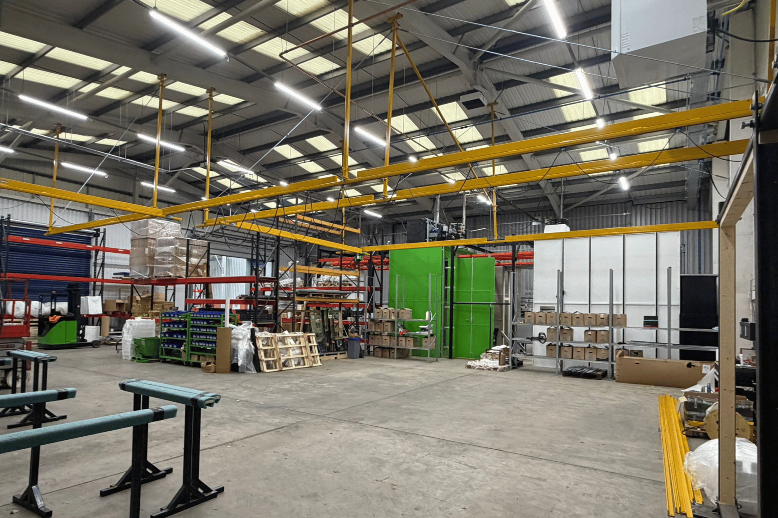 The image shows the interior of a warehouse or factory with high ceilings, industrial lighting, and various equipment and supplies stacked on shelves and the floor. There is yellow overhead crane equipment and green partition walls visible, along with pallets and packaging materials.