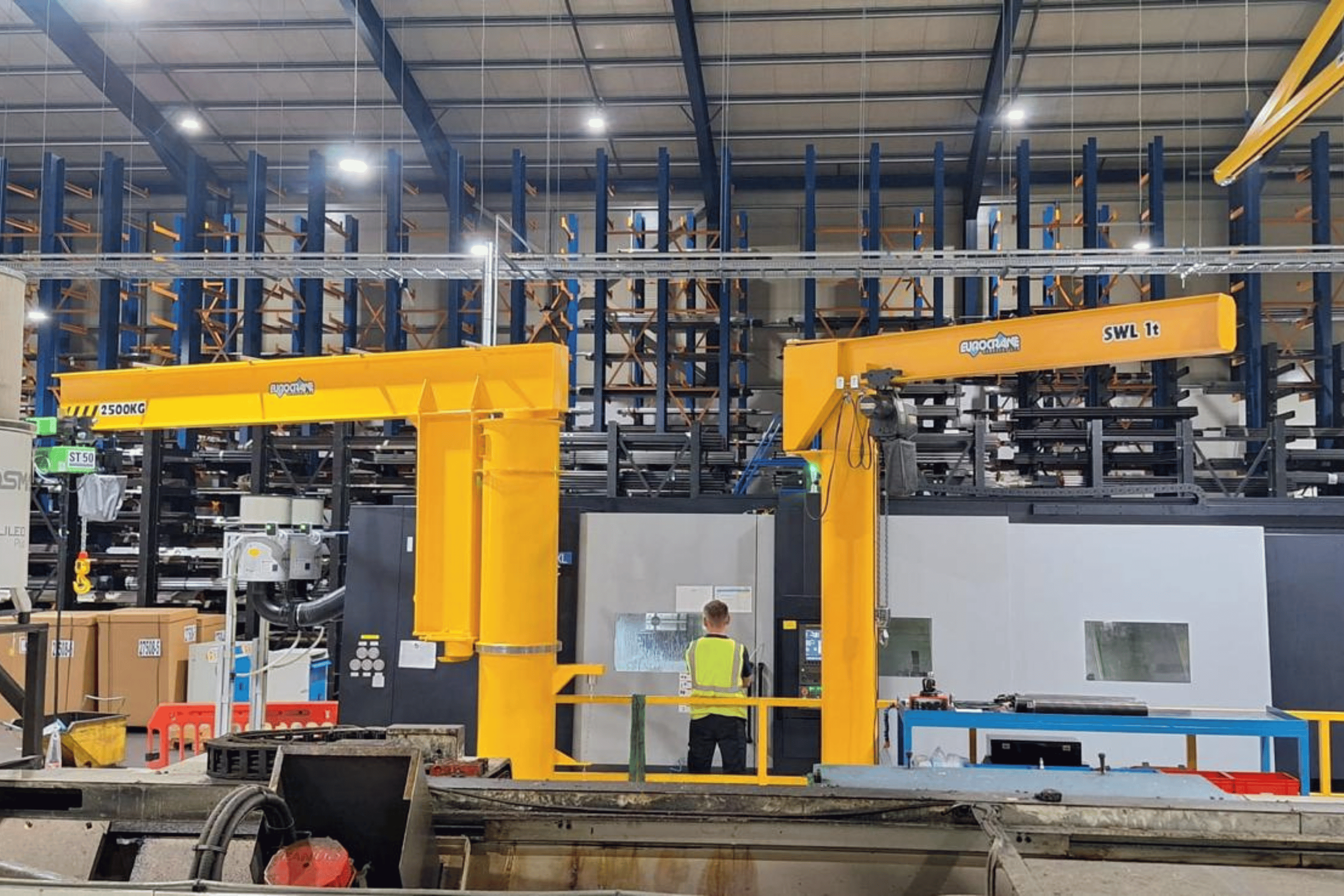 Inside a warehouse with tall blue shelving and orange support beams. Large yellow cranes with weight capacities are present. A person wearing a yellow vest stands in front of the equipment. Various boxes and equipment are visible.