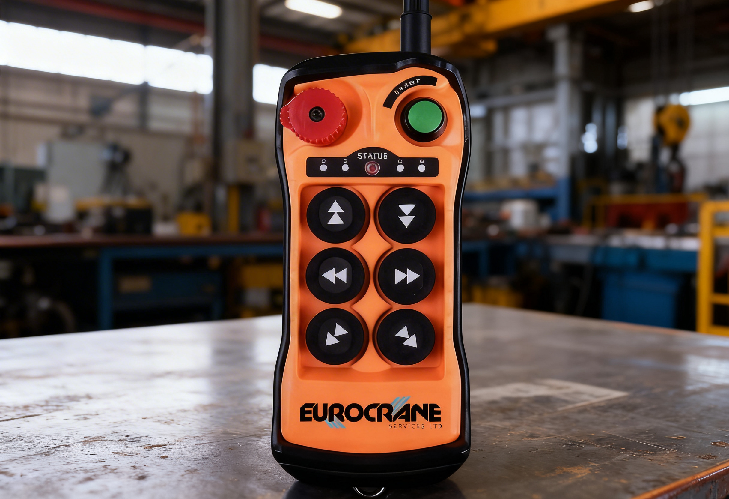Orange remote control for Eurocrane services, featuring multiple black and white buttons, a red dial, a green button, and labeled controls, placed on a work surface in a warehouse or industrial setting.