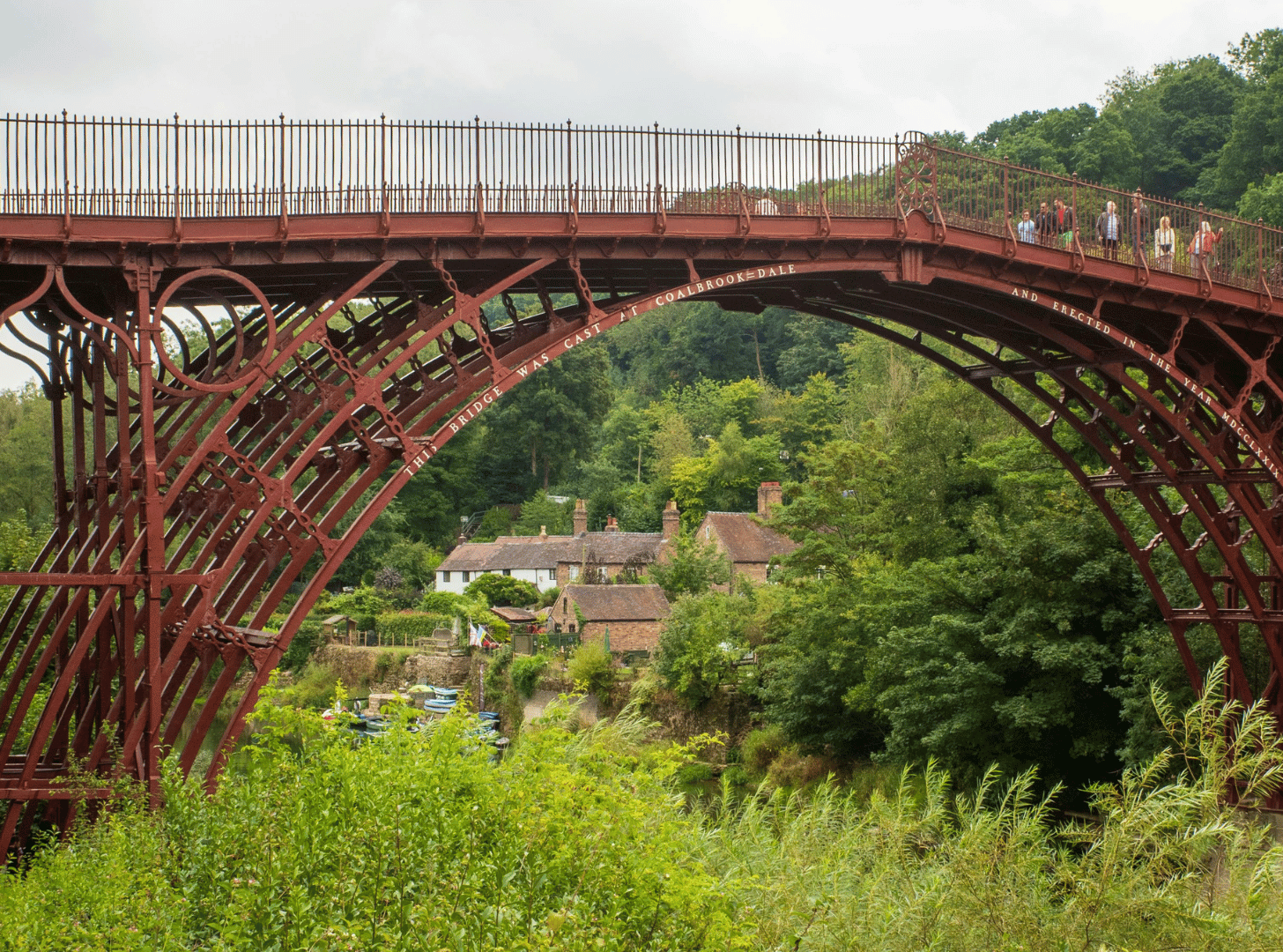The Clifton Suspension Bridge spans across a lush green valley with houses and trees below, and people walking on the bridge.