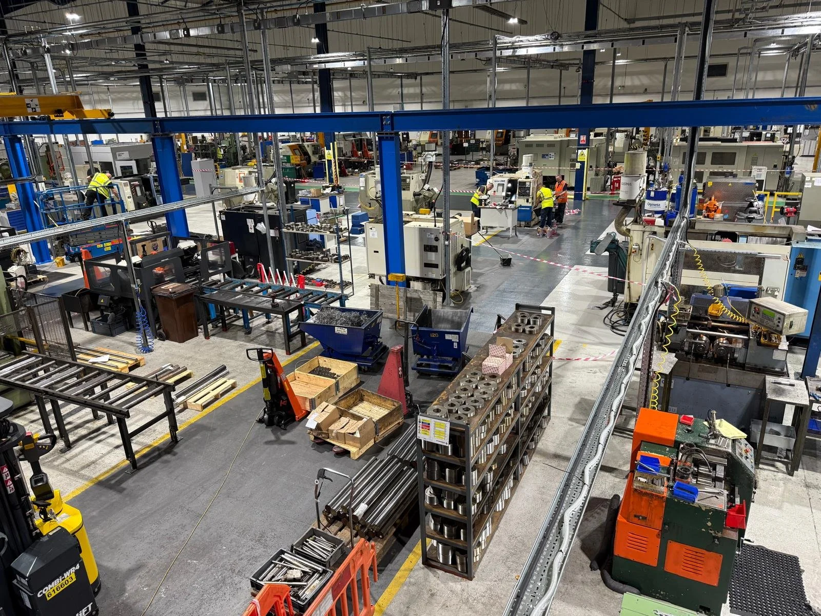 View of a busy manufacturing or industrial workshop with workers in safety vests operating machinery, metal shelves, and various equipment and tools, with a yellow obstacle in the foreground.