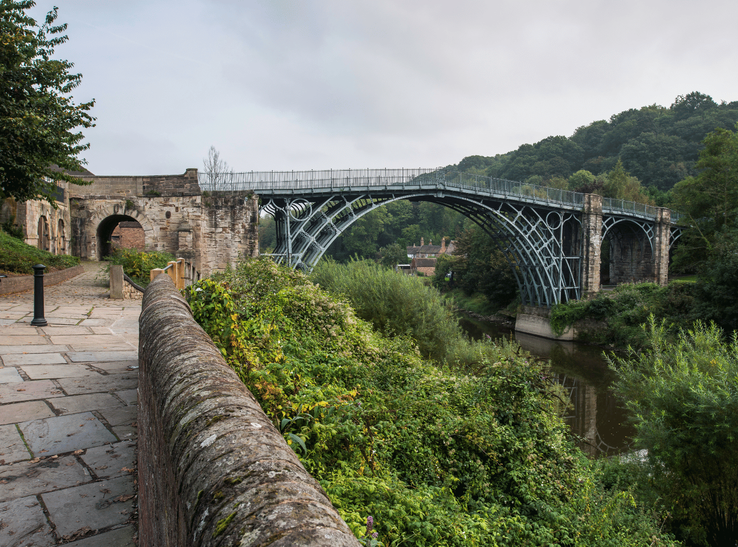 An old stone bridge with metal support structure spans over a river, with lush greenery on both sides and a cloudy sky overhead.