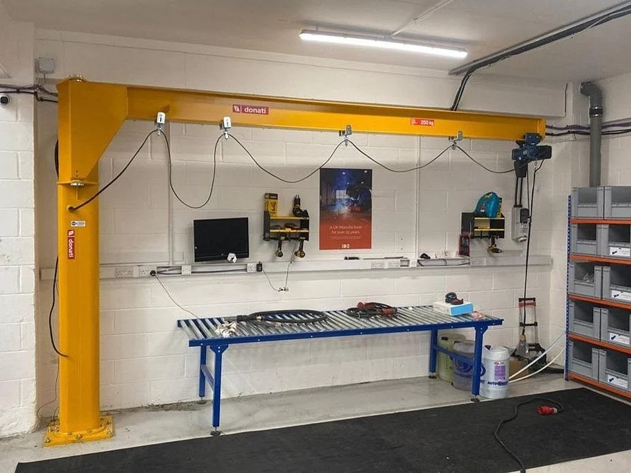 Workshop with a yellow overhead crane labeled 'donati' and '250 kg', a workbench with tools and a black monitor, wall-mounted shelves with tools, storage bins, and electrical outlets, and a black rubber mat on the floor.