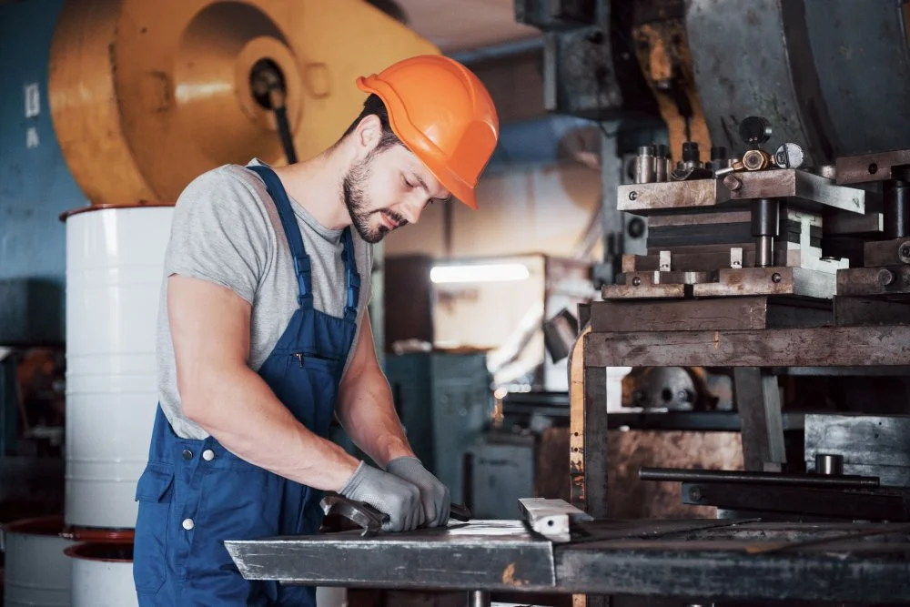 A man wearing a gray shirt, blue overalls, gray gloves, and an orange safety helmet working with metal on a workbench in an industrial workshop.