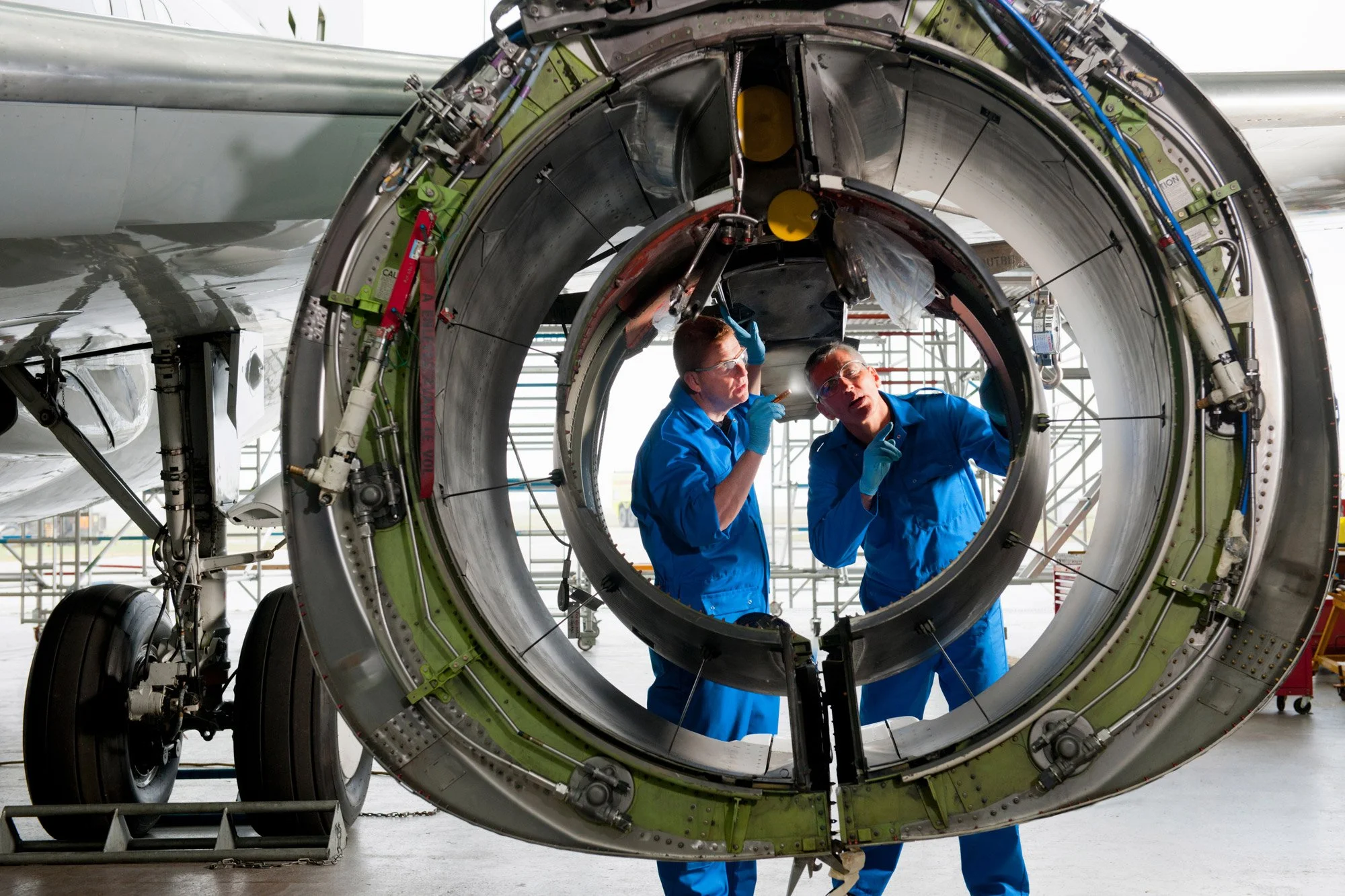 Two technicians in blue coveralls and safety glasses are inspecting an aircraft engine removed from the aircraft, inside a hangar, with scaffolding in the background.