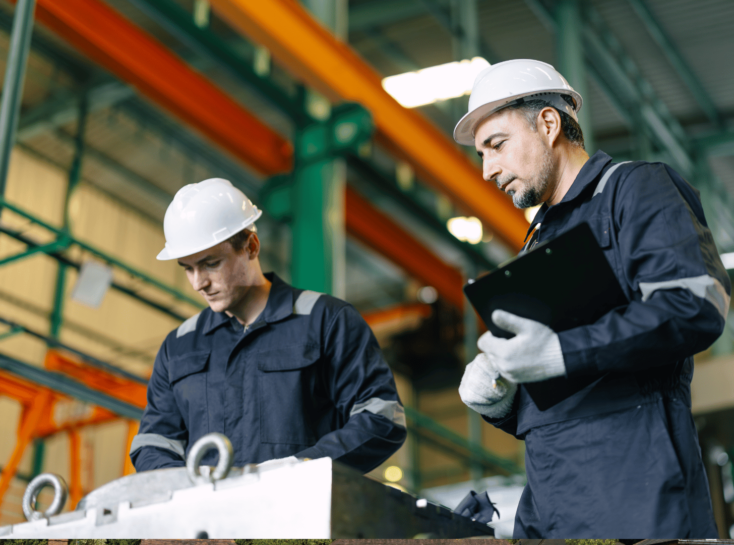 Two male workers in safety helmets and uniforms inspecting machinery in an industrial warehouse.