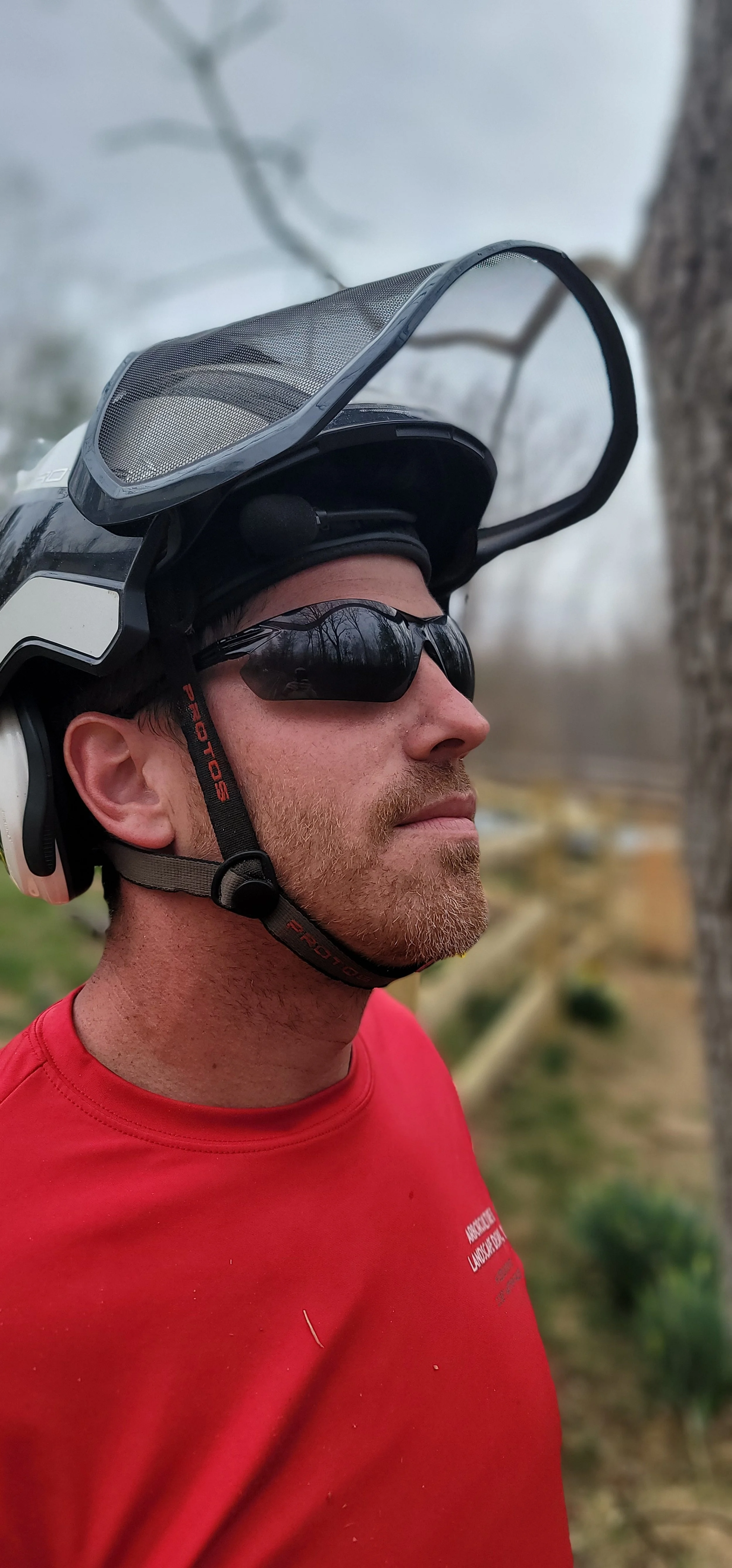 A man wearing sunglasses, a red shirt, and a helmet with a face shield attached, standing outdoors near a tree.