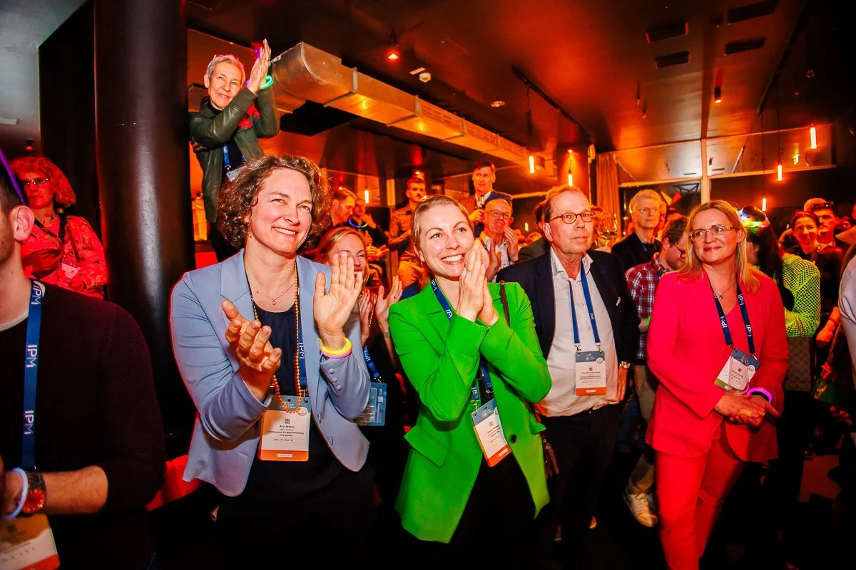 Menschen bei einer Veranstaltung, applaudierend und im Publikum sitzend, in einem dunklen Raum mit rot beleuchteter Decke.