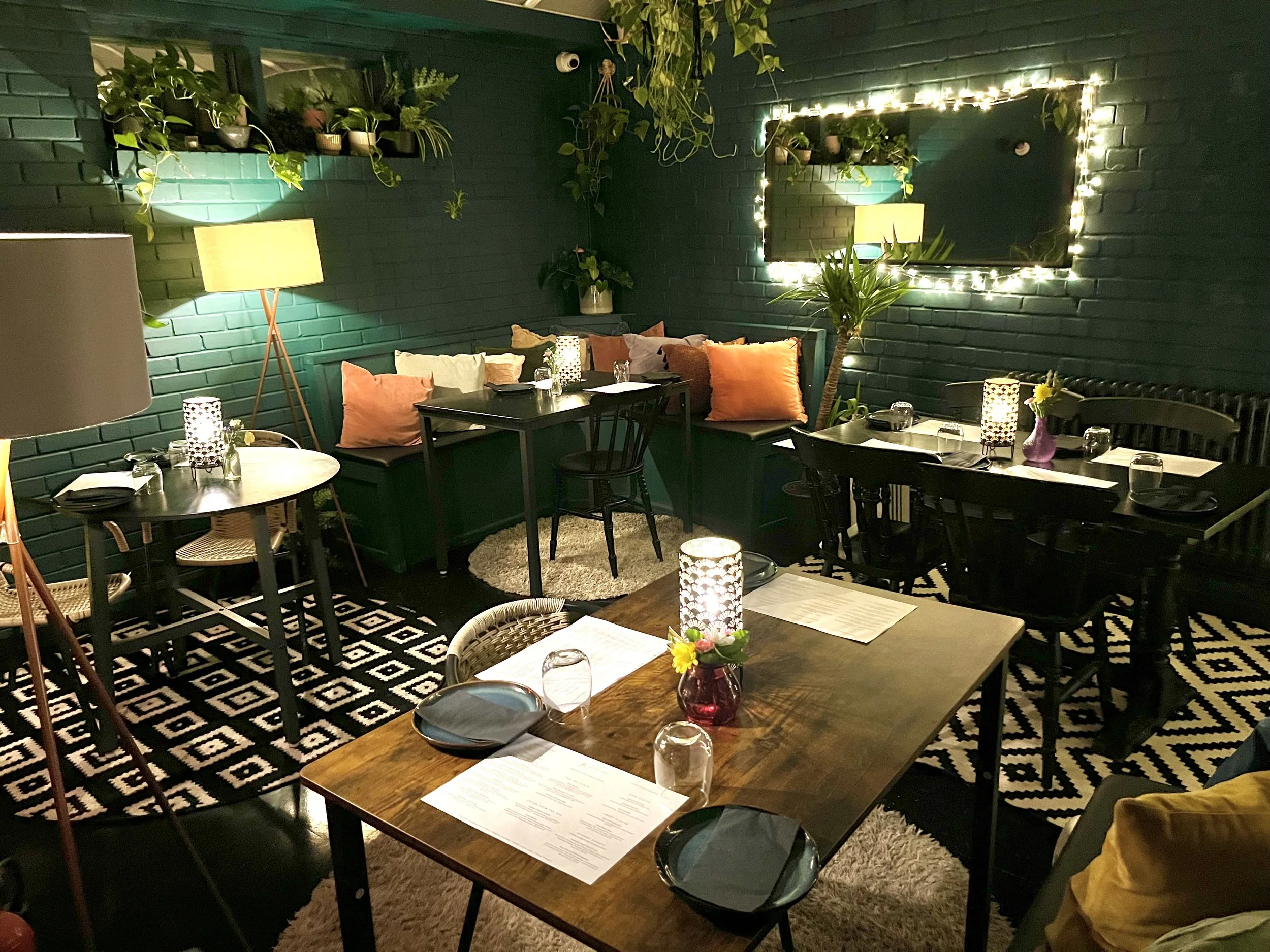 Interior of a cozy restaurant with dark green brick walls, decorated with hanging plants, string lights, and a mirror. There are various tables set with black plates, glasses, and candles, with colorful cushions and a checkered rug.