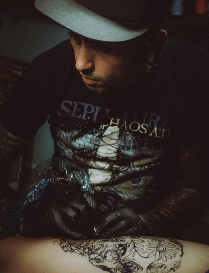 A man with a hat tattooing a floral design on someone's arm.