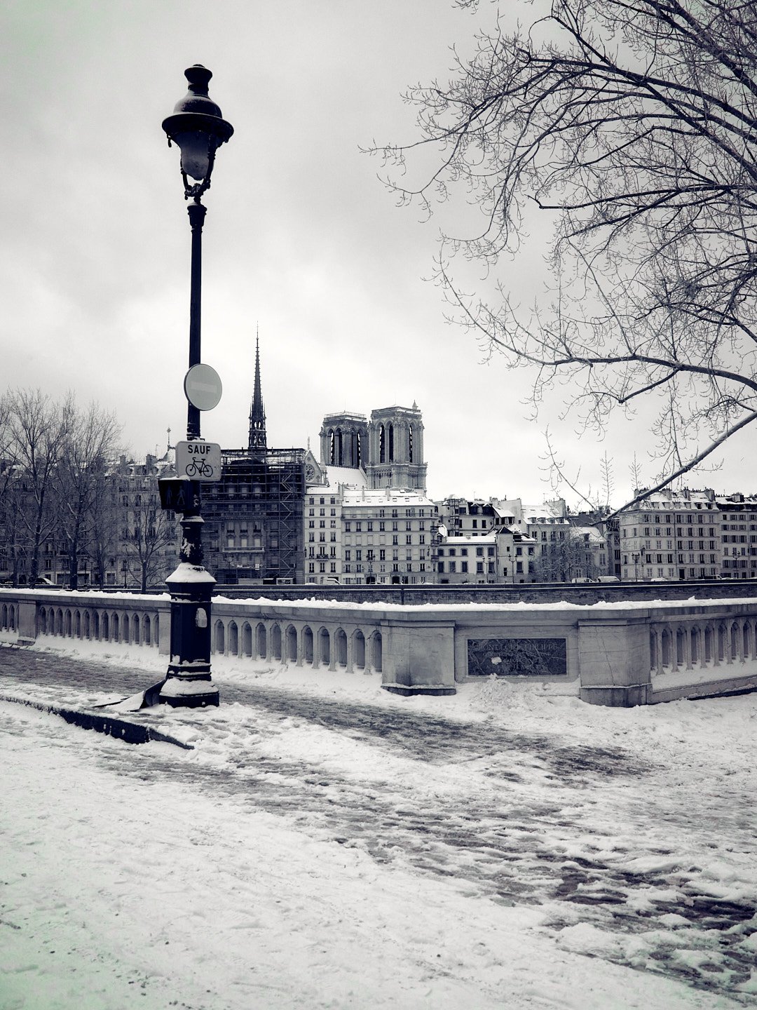 Snow-covered bridge with street lamp in front of historic Parisian buildings and Notre-Dame Cathedral in the distance on a gray winter day.