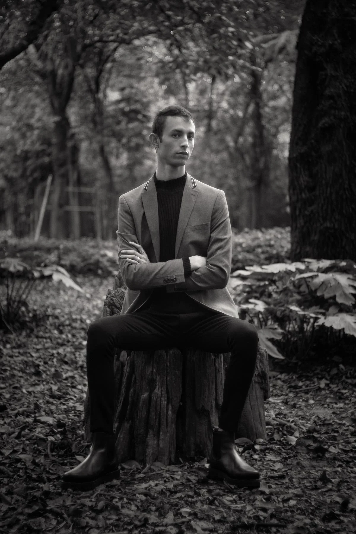 A young man in a suit sitting on a tree stump in a forest, looking to the side, in black and white. He has his arms crossed and is wearing black boots.