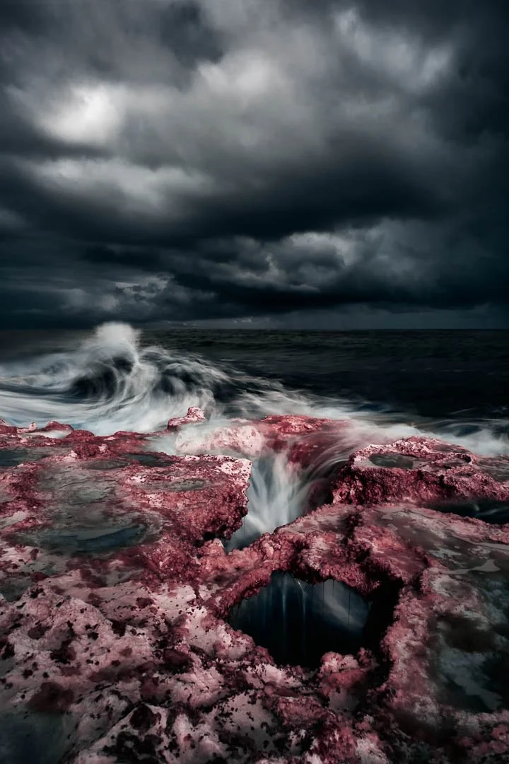 Stormy ocean scene with dark clouds, choppy waves, and pinkish rocks in the foreground