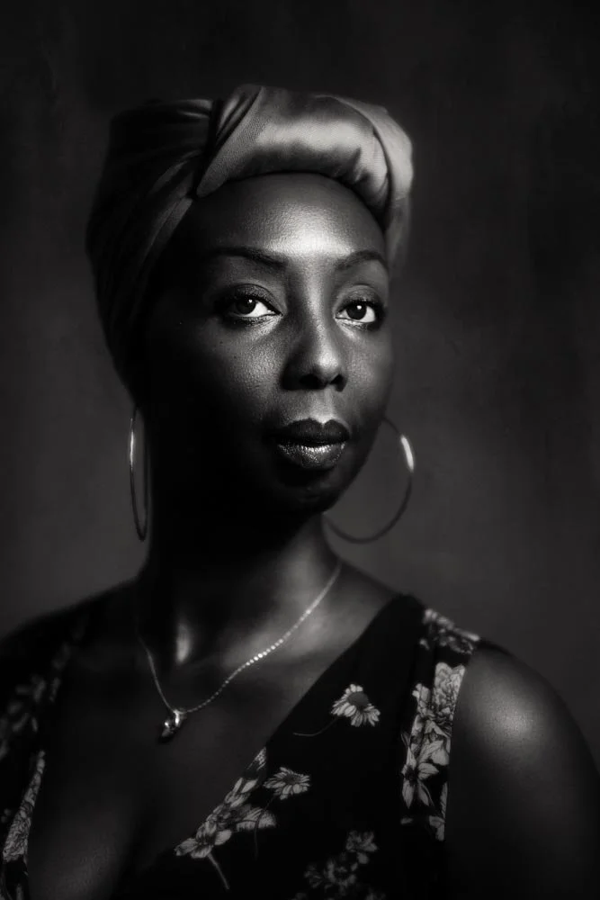 Black and white portrait of a woman wearing a head wrap, hoop earrings, and a floral dress, with a necklace, looking poised and elegant.
