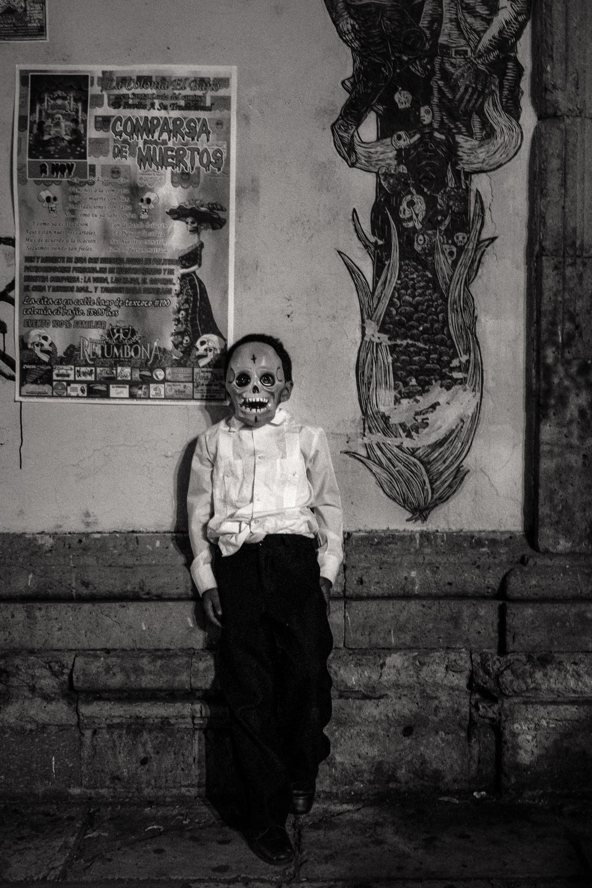 Child dressed in a skeleton masquerade mask and formal attire leaning against a wall with a large Frida Kahlo-inspired mural and a poster in the background.