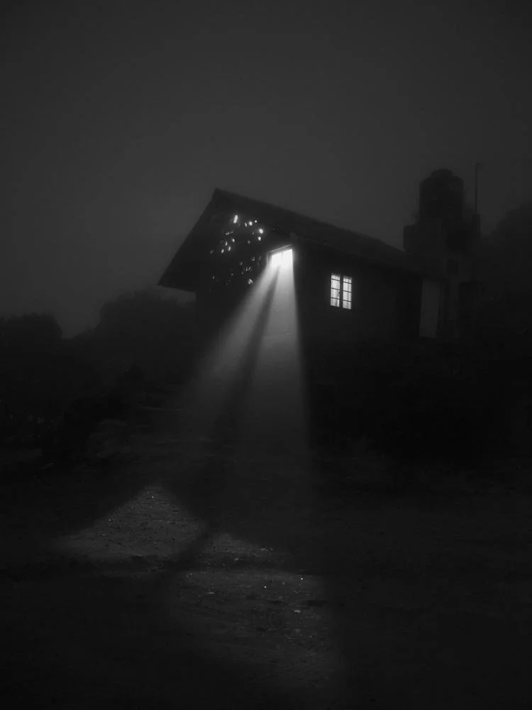 A dark, foggy night scene with a tilted barn that has holes in its roof. Light is shining through the holes and a window, illuminating the ground with beams of light.