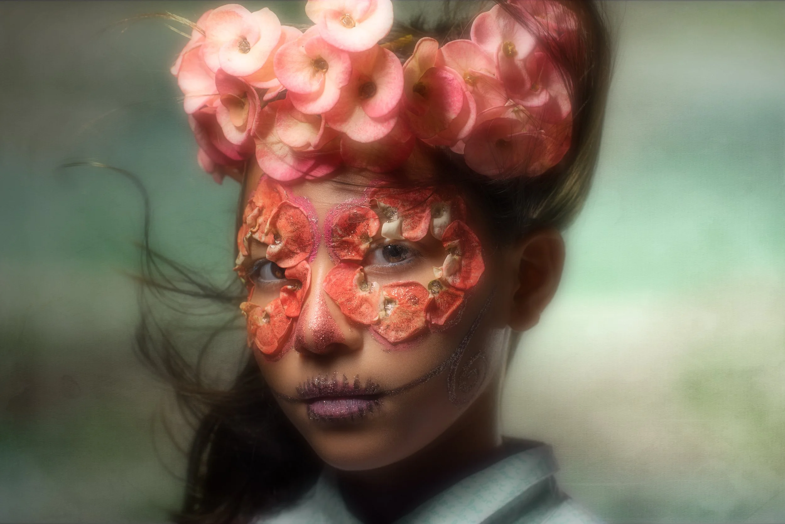 A person with face paint resembling a sugar skull, featuring pink and purple hues, with flowers on top of their head and petals arranged over their eyes and cheeks.