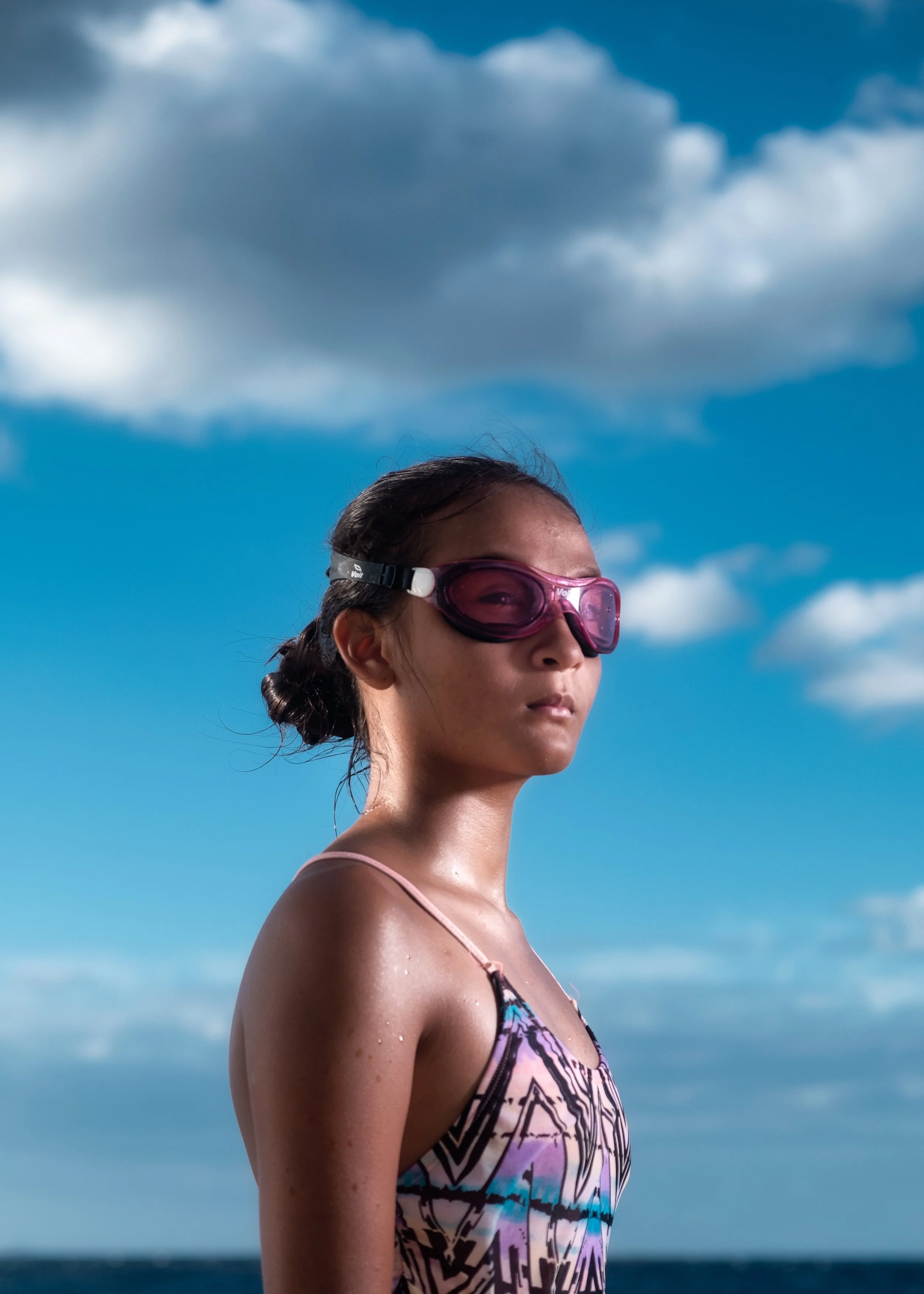 A young girl with sunglasses standing outdoors under a cloudy sky, wearing a colorful patterned swimsuit.