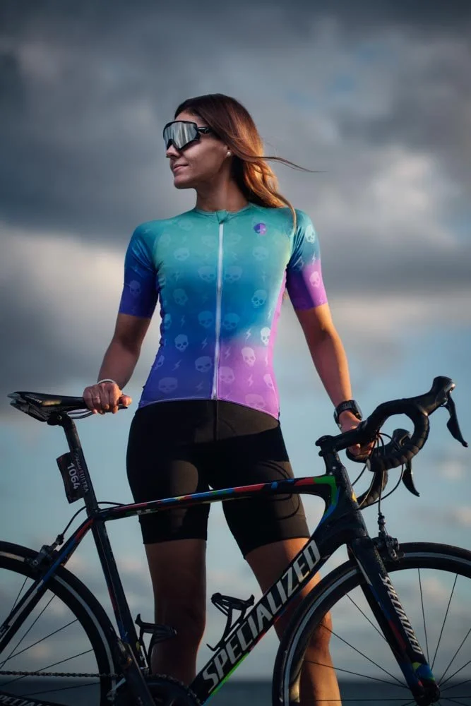 A woman in cycling gear, wearing sunglasses and a colorful skull-patterned jersey, stands outdoors with a black Specialized road bike against a cloudy sky.
