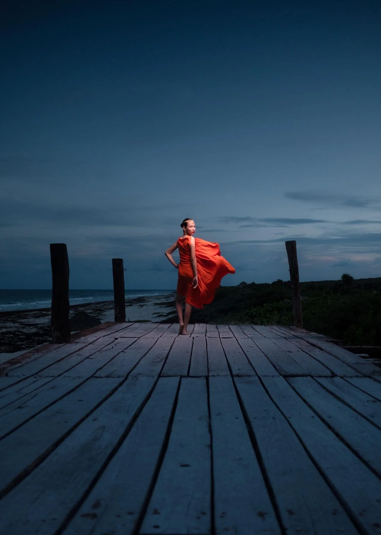 A woman in an orange dress standing on a wooden pier at dusk, overlooking the ocean with a cloudy sky.