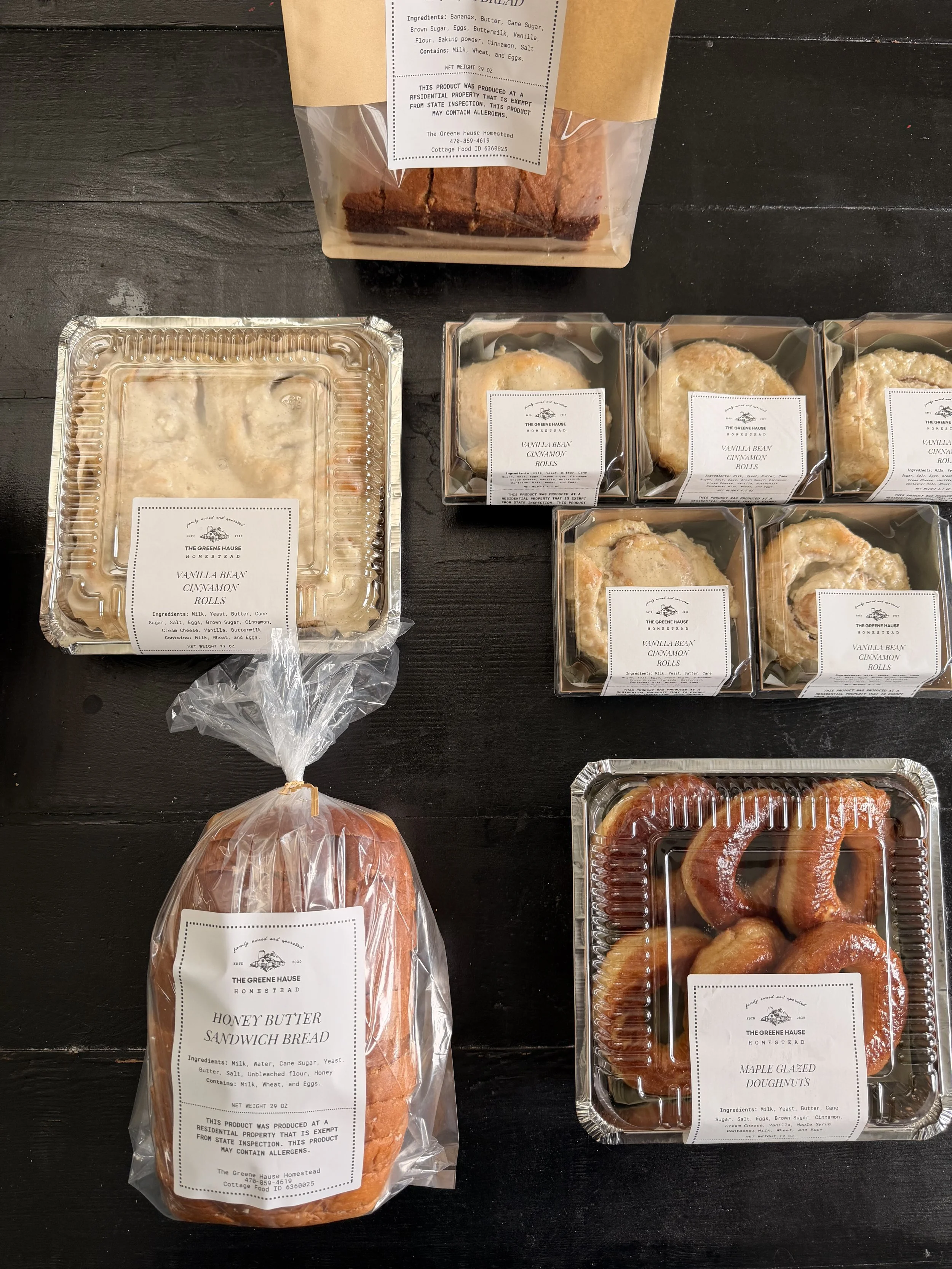 Various packaged baked goods, including vanilla bean cinnamon rolls, honey butter sandwich bread, maple glazed doughnuts, and slices of bread, displayed on a dark surface.
