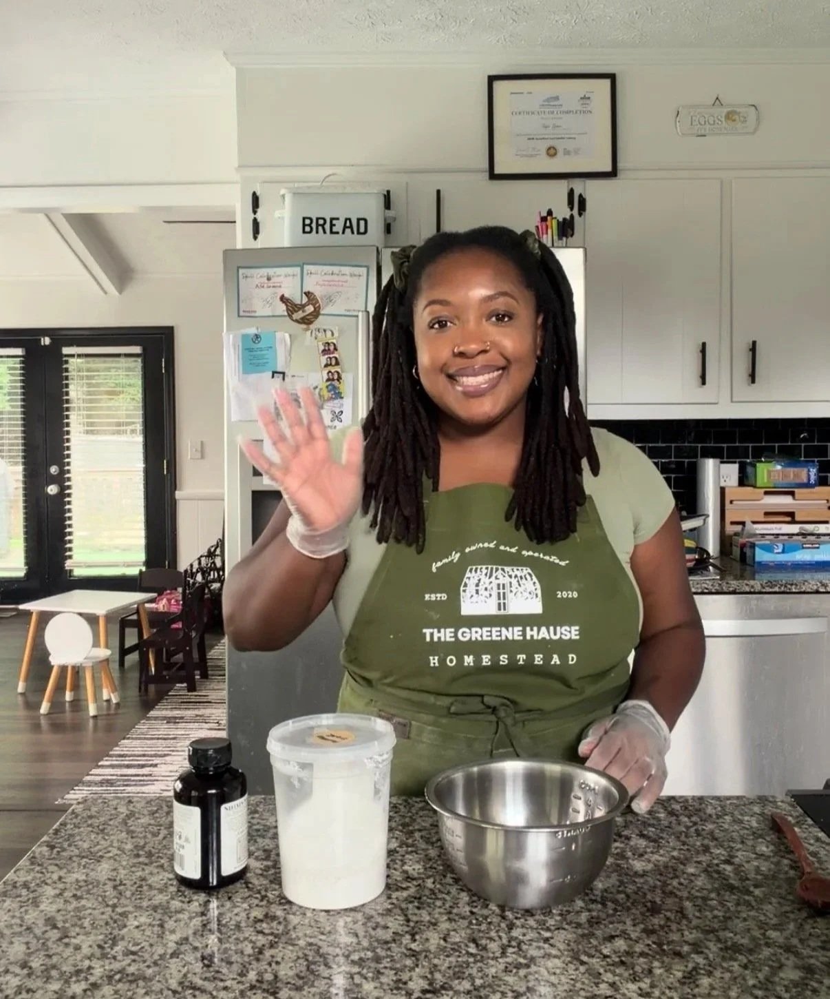 A woman wearing a green apron and a glove on her left hand, smiling and waving with her right hand, standing in a kitchen with a bowl and ingredients on the counter.