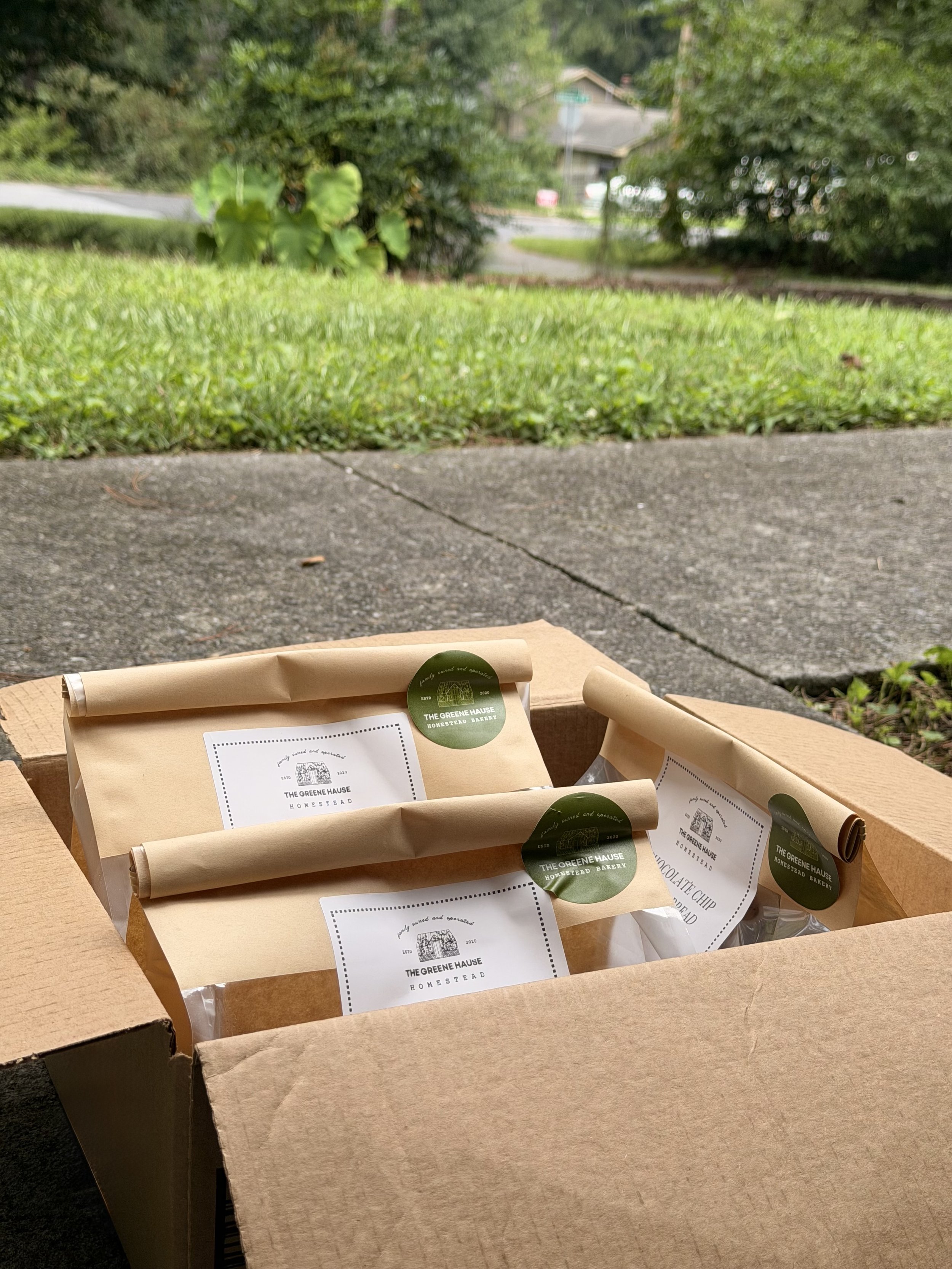 Open cardboard box containing wrapped baked goods labeled from The Greene Hause Homestead Bakery, placed on a wooden surface outdoors with house and trees in the background.