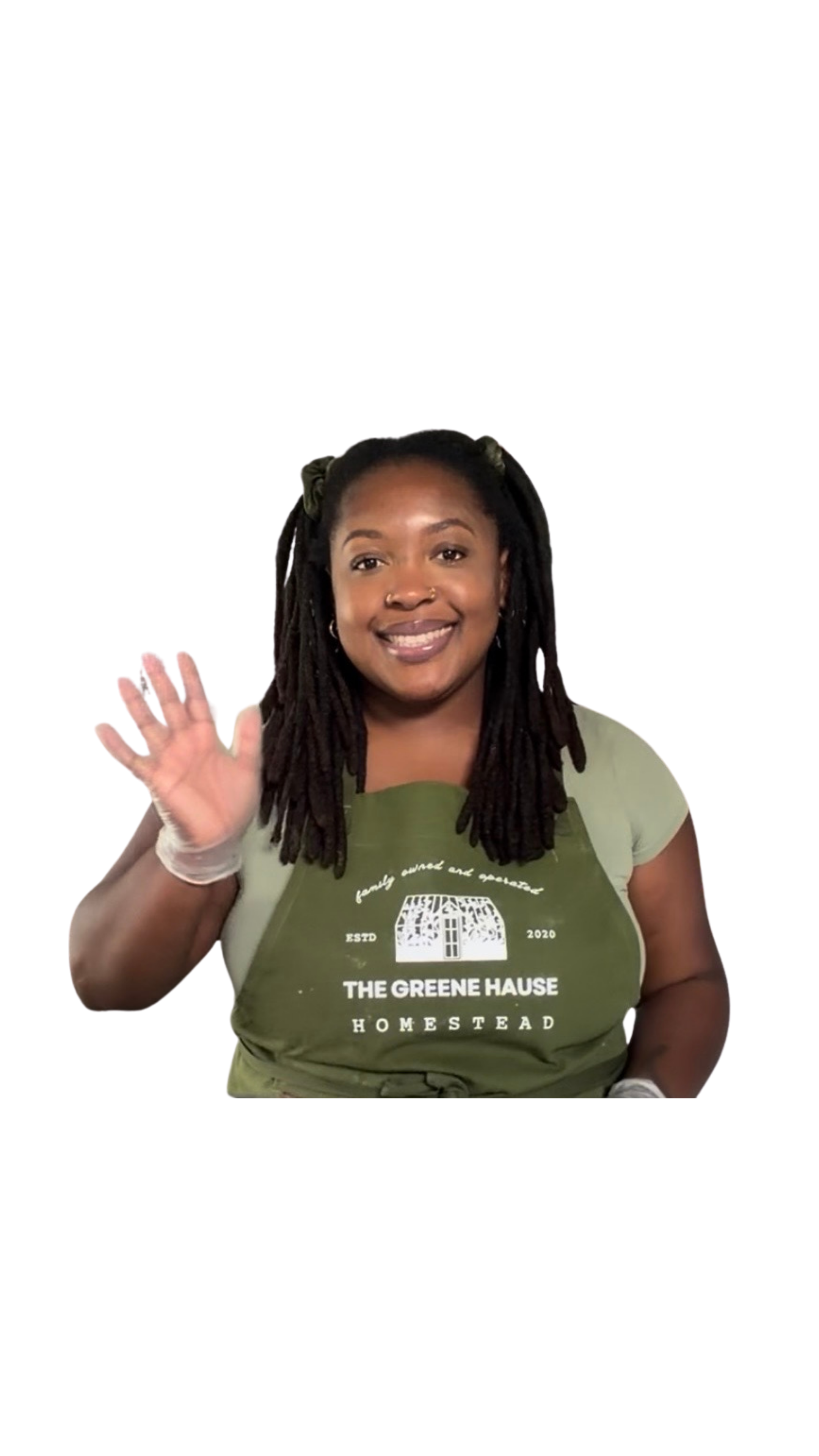 A smiling woman with long dreadlocks waving, wearing a green T-shirt with white text and a green apron that reads "The Greene Hauge Homestead".