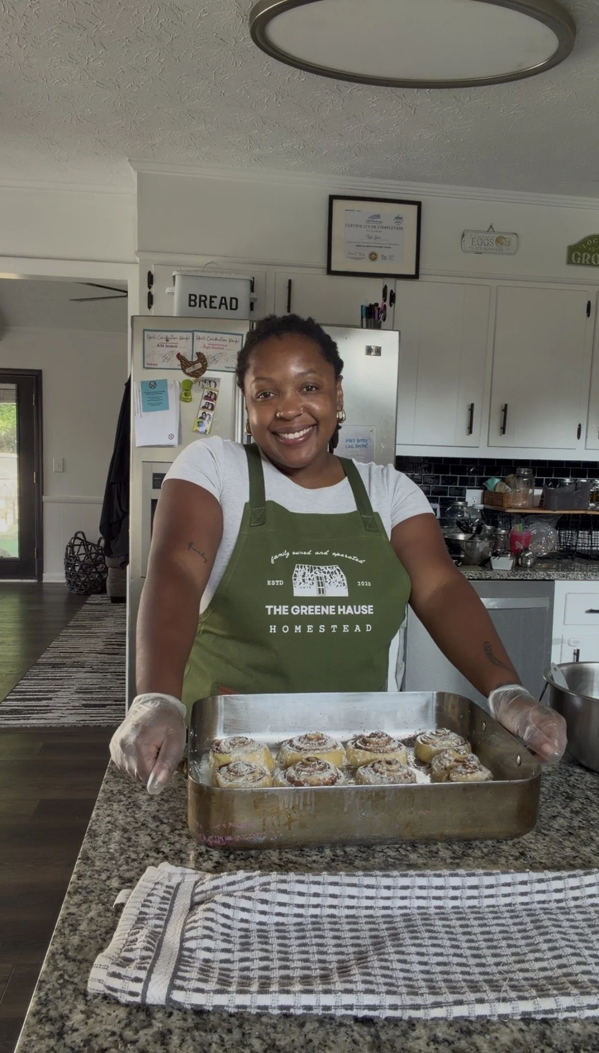 A woman smiling at the camera while wearing a green apron and white gloves, holding a baking tray of cinnamon rolls in a kitchen with white cabinets and a countertop.