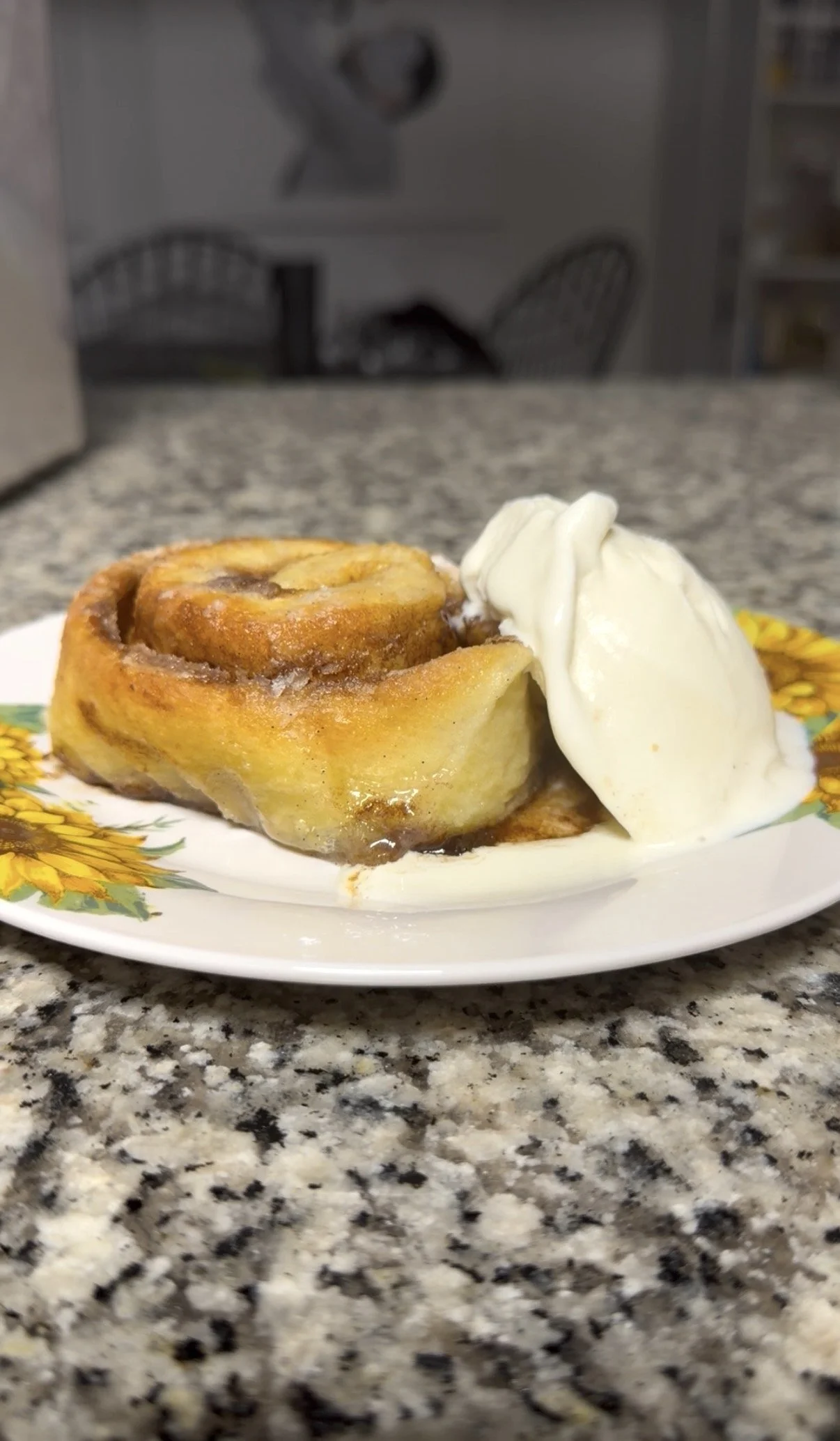 A plate with a cinnamon roll and a scoop of vanilla ice cream on a granite countertop, with a blurred kitchen background.