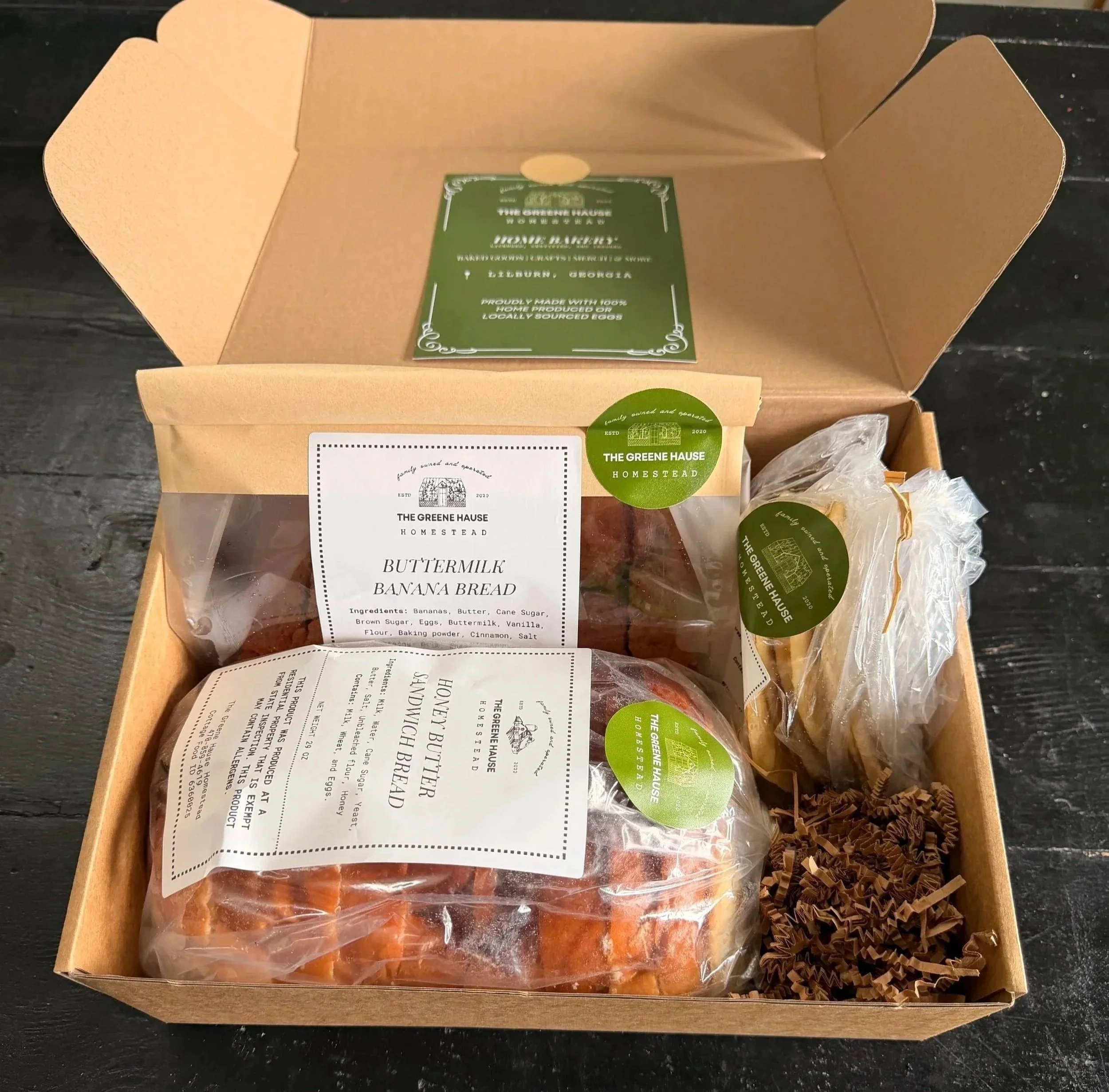 Open cardboard box containing a package of buttermilk banana bread from The Greene Haus Homestead, with a green label and a white label with product details.