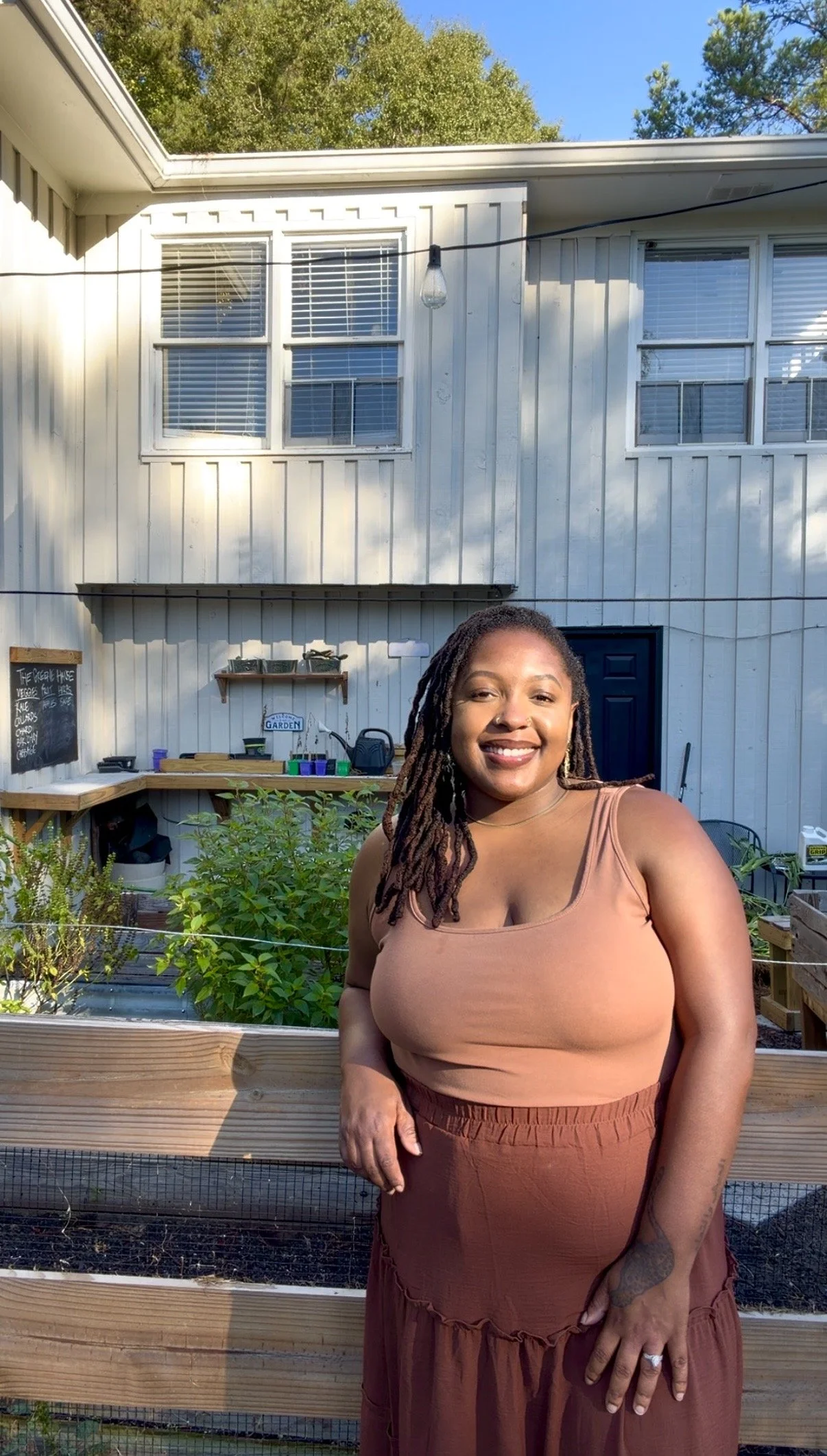 A woman with dreadlocks and a brown tank top smiling outdoors in a backyard garden with a wooden raised bed and green plants, against a white house with multiple windows and string lights.