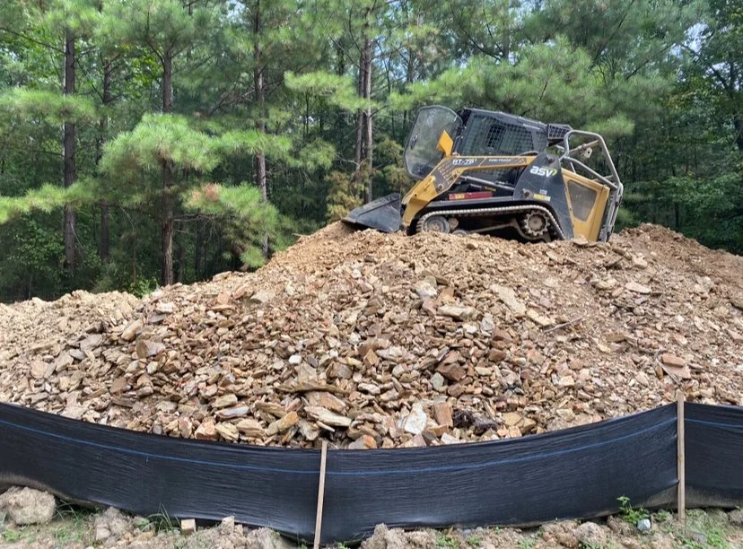 A small bulldozer on a pile of dirt and rocks, with a wooded forest in the background.