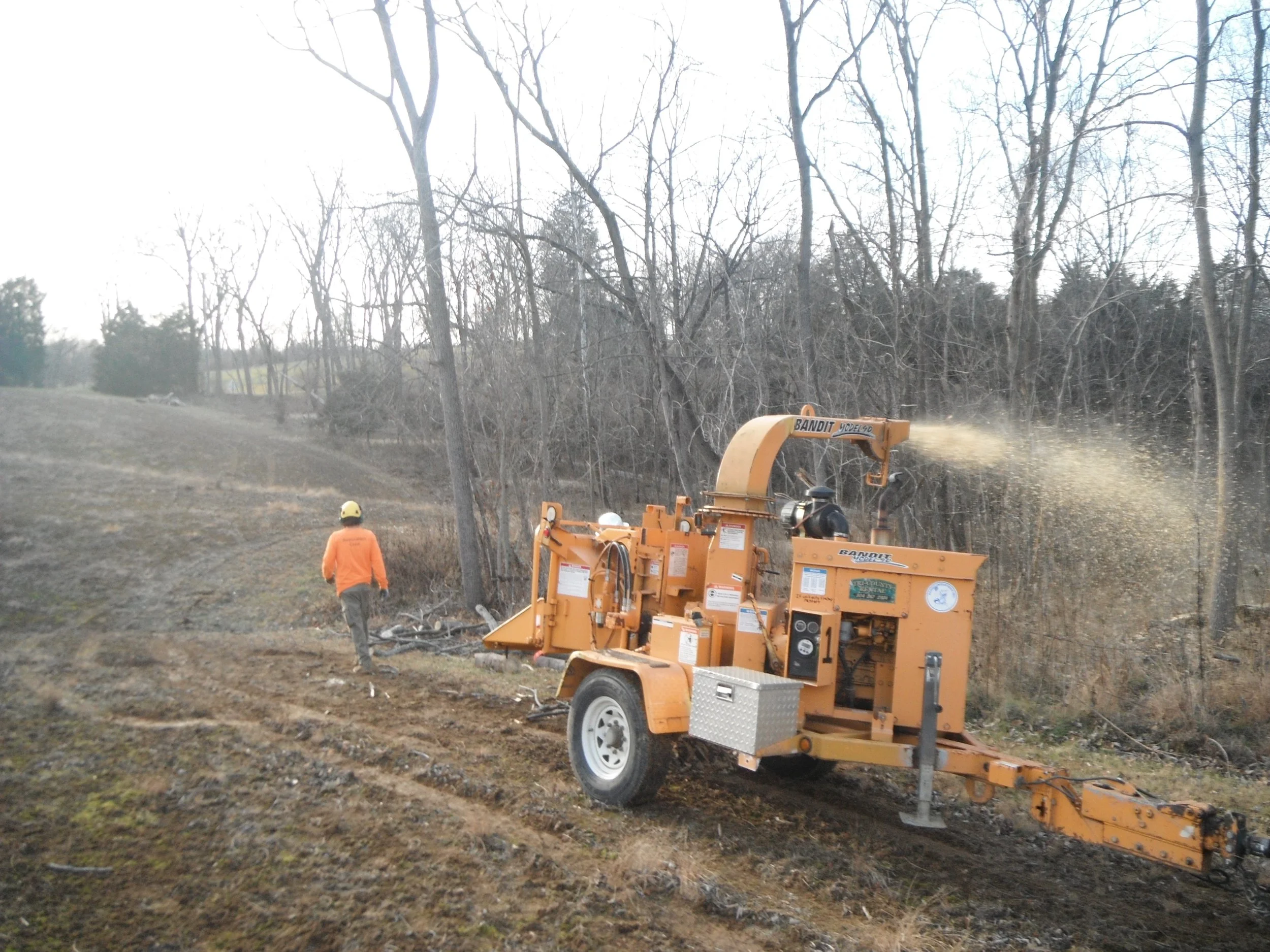 A forestry contractor wearing an orange shirt and yellow helmet walking next to a wood chipper on a grassy and dirt area, with trees in the background.