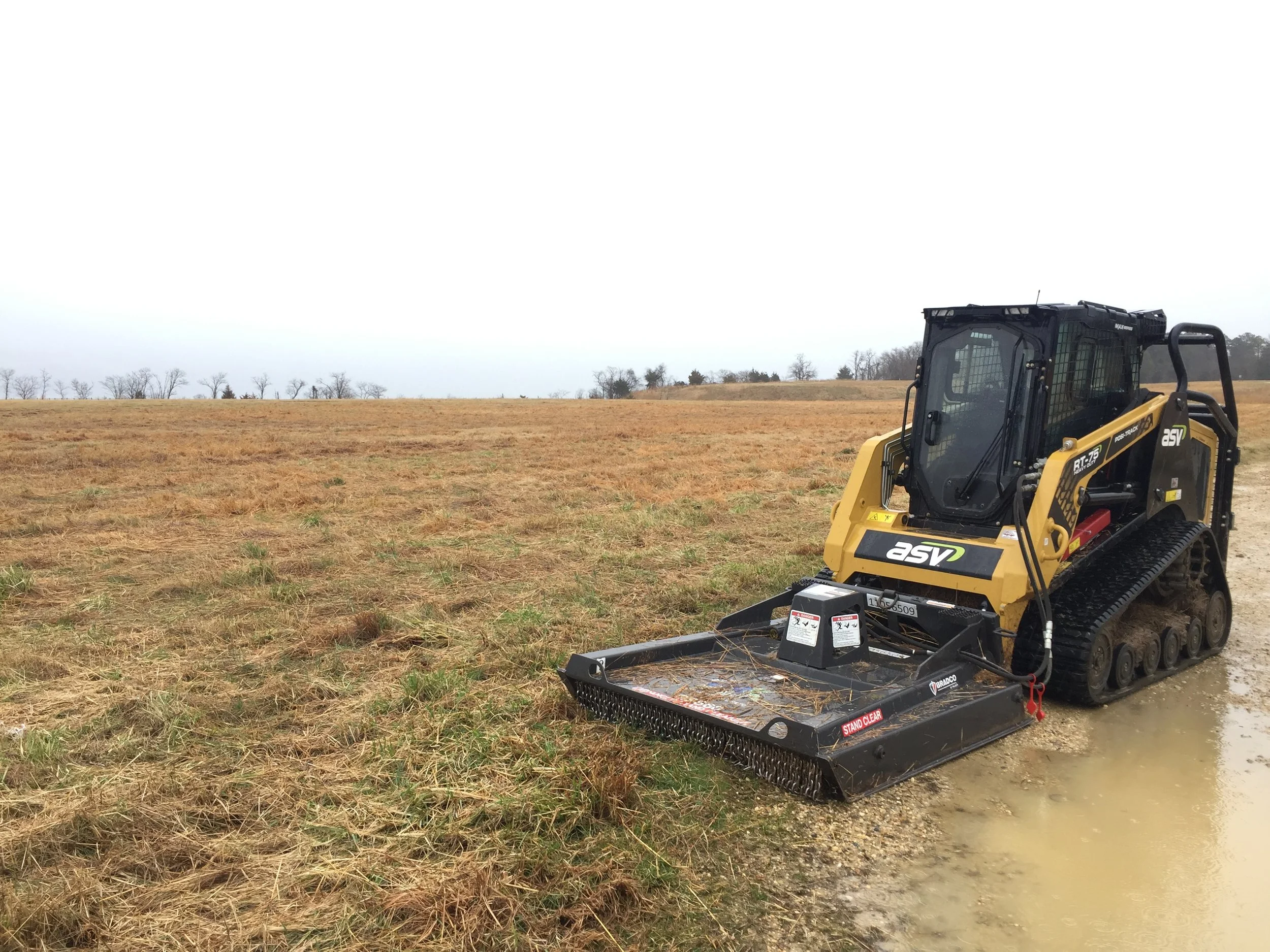 A small tracked skid steer loader on a muddy field with droughted grass and a cloudy sky in the background.
